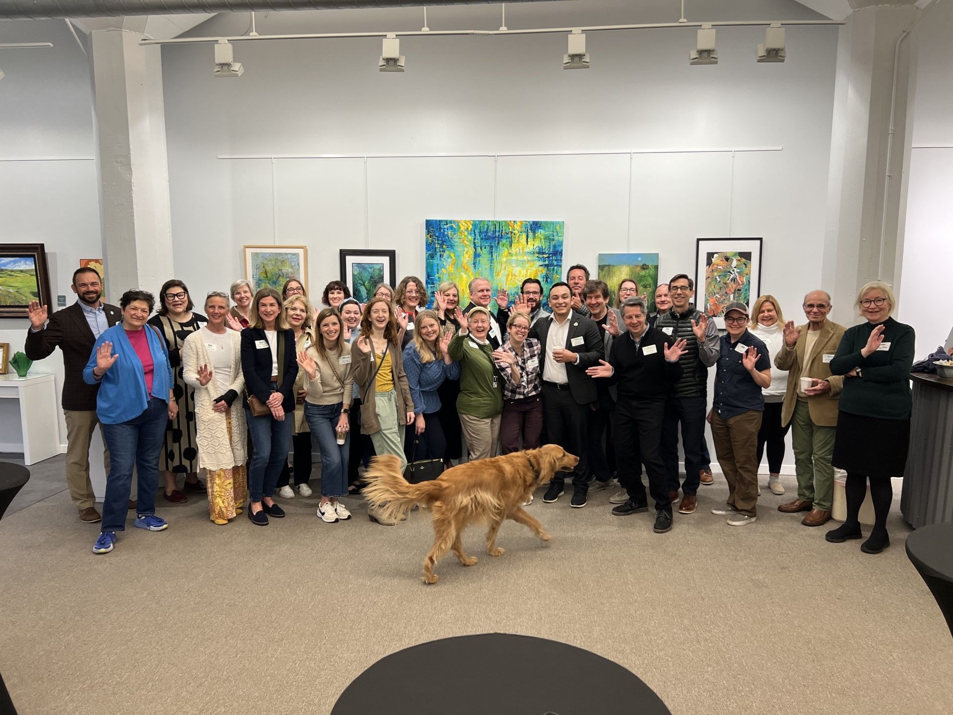Large group poses in an art gallery, smiling and waving. A golden retriever dog walks toward the center.