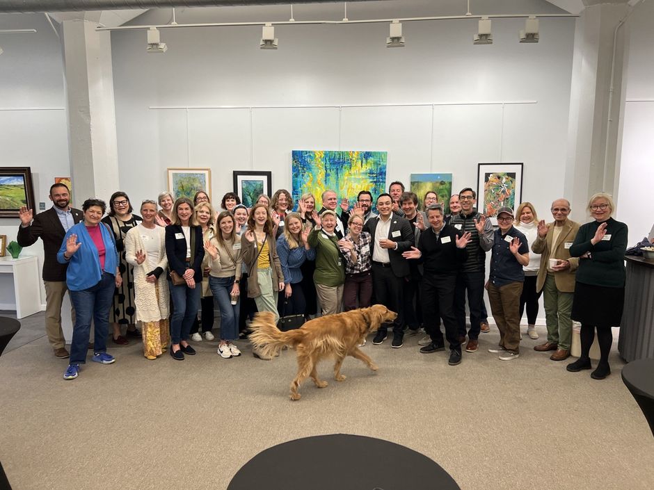 Large group poses in an art gallery, smiling and waving. A golden retriever dog walks toward the center.