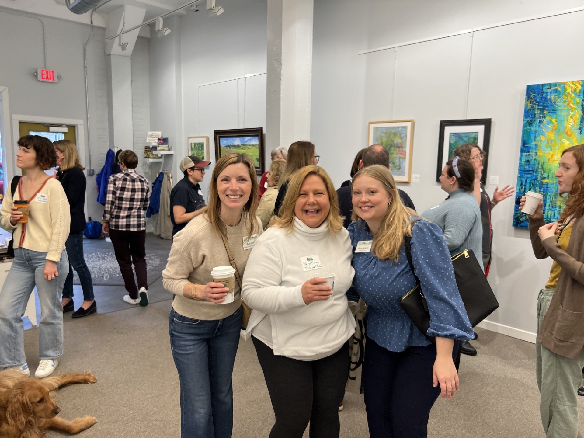 Three women smiling at the camera in a gallery setting, holding drinks. Other people and art in the background.