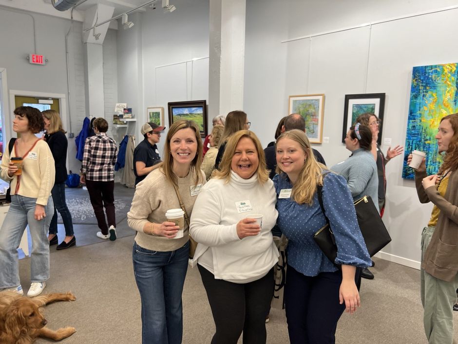 Three women smiling at the camera in a gallery setting, holding drinks. Other people and art in the background.