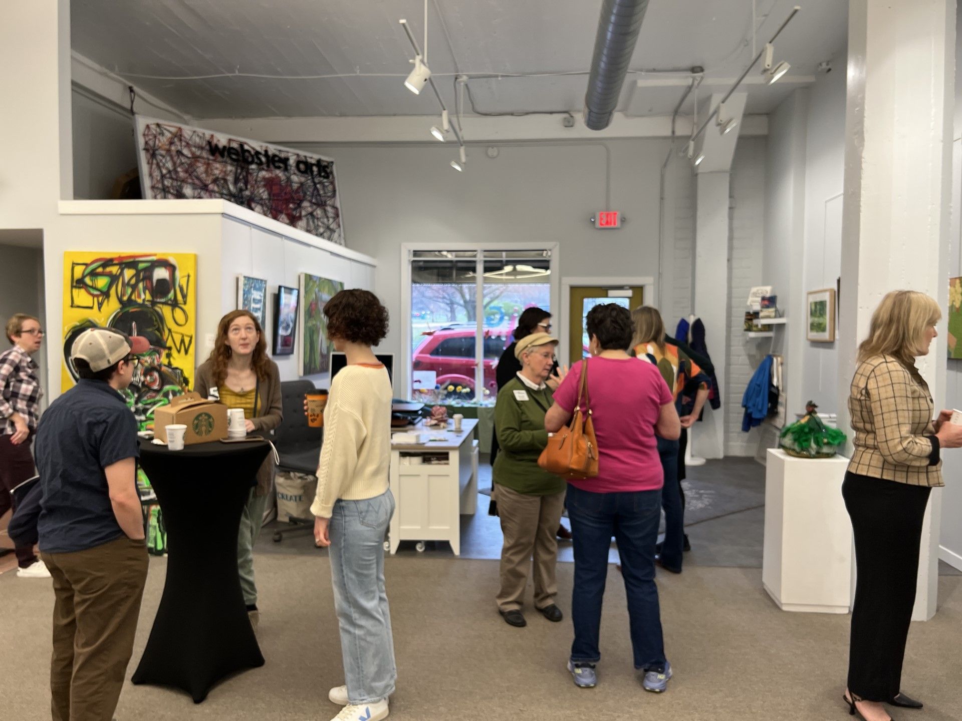 People at an art gallery opening, viewing paintings and socializing. White walls, various artworks, and drinks.