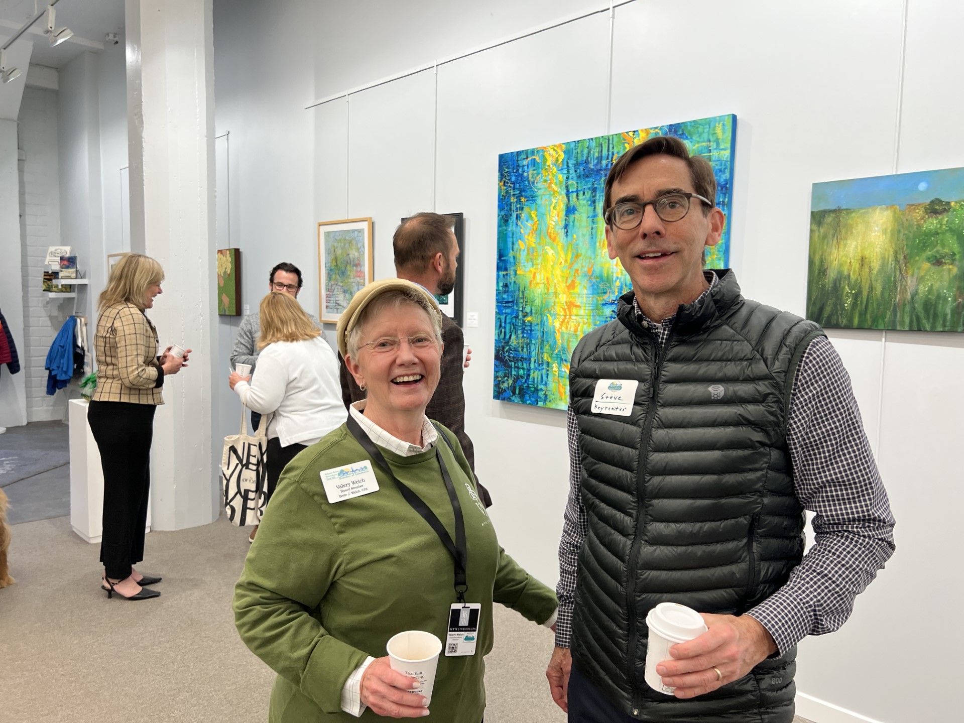 Two people smiling at an art gallery, holding cups. Paintings on the walls, other visitors in the background.