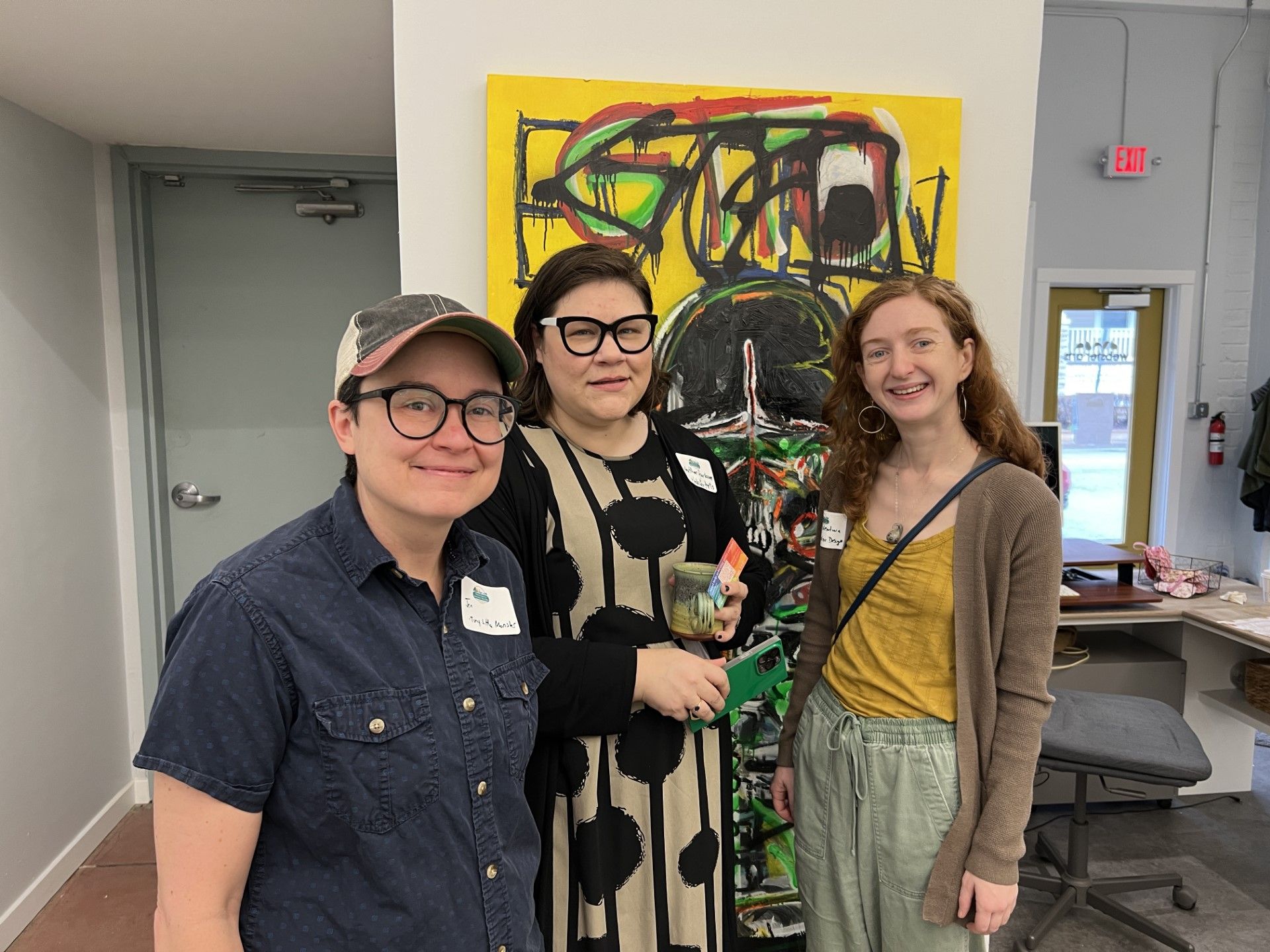 Three people stand in front of a colorful abstract painting. One wears glasses, a denim shirt, and a hat; another has glasses and a patterned dress; the third has a cardigan.