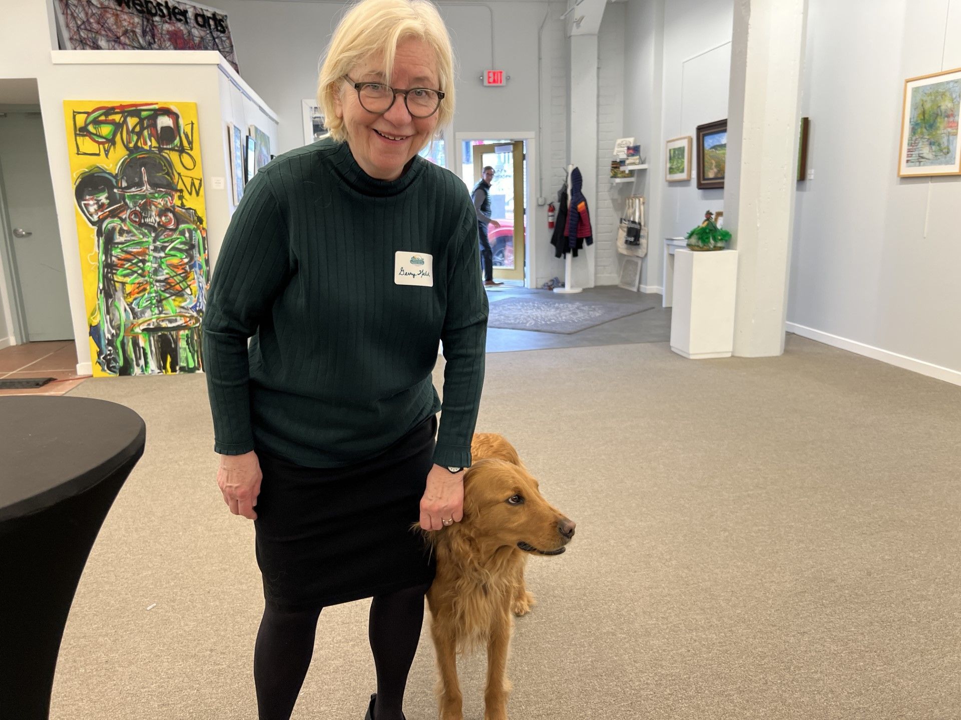 Woman and dog in art gallery, woman smiling, dog looking forward.