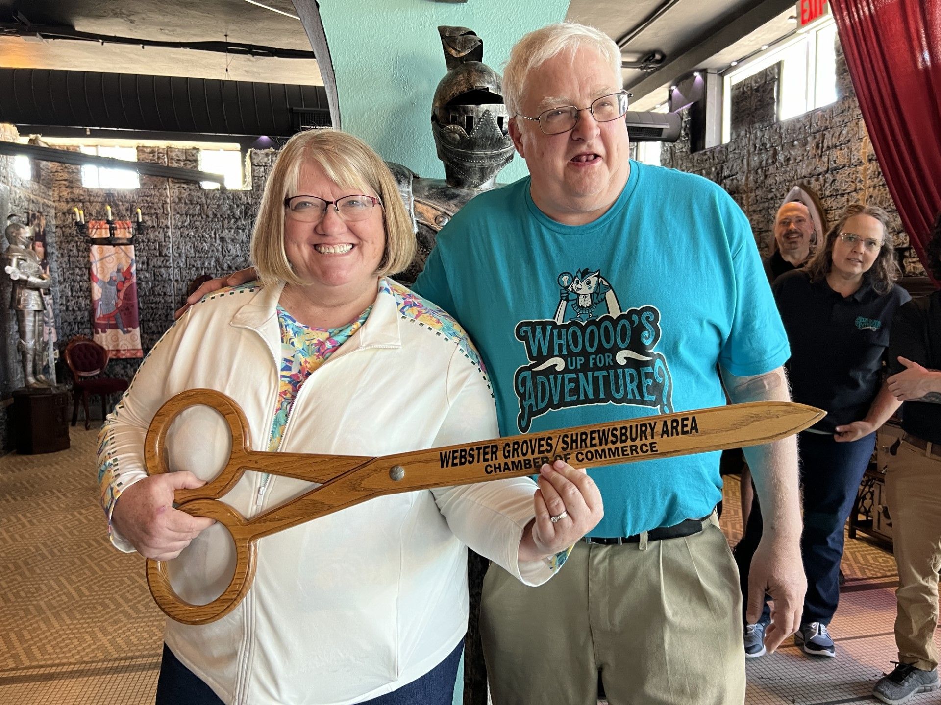 Couple holding giant scissors at an event; indoor setting with onlookers and artwork.