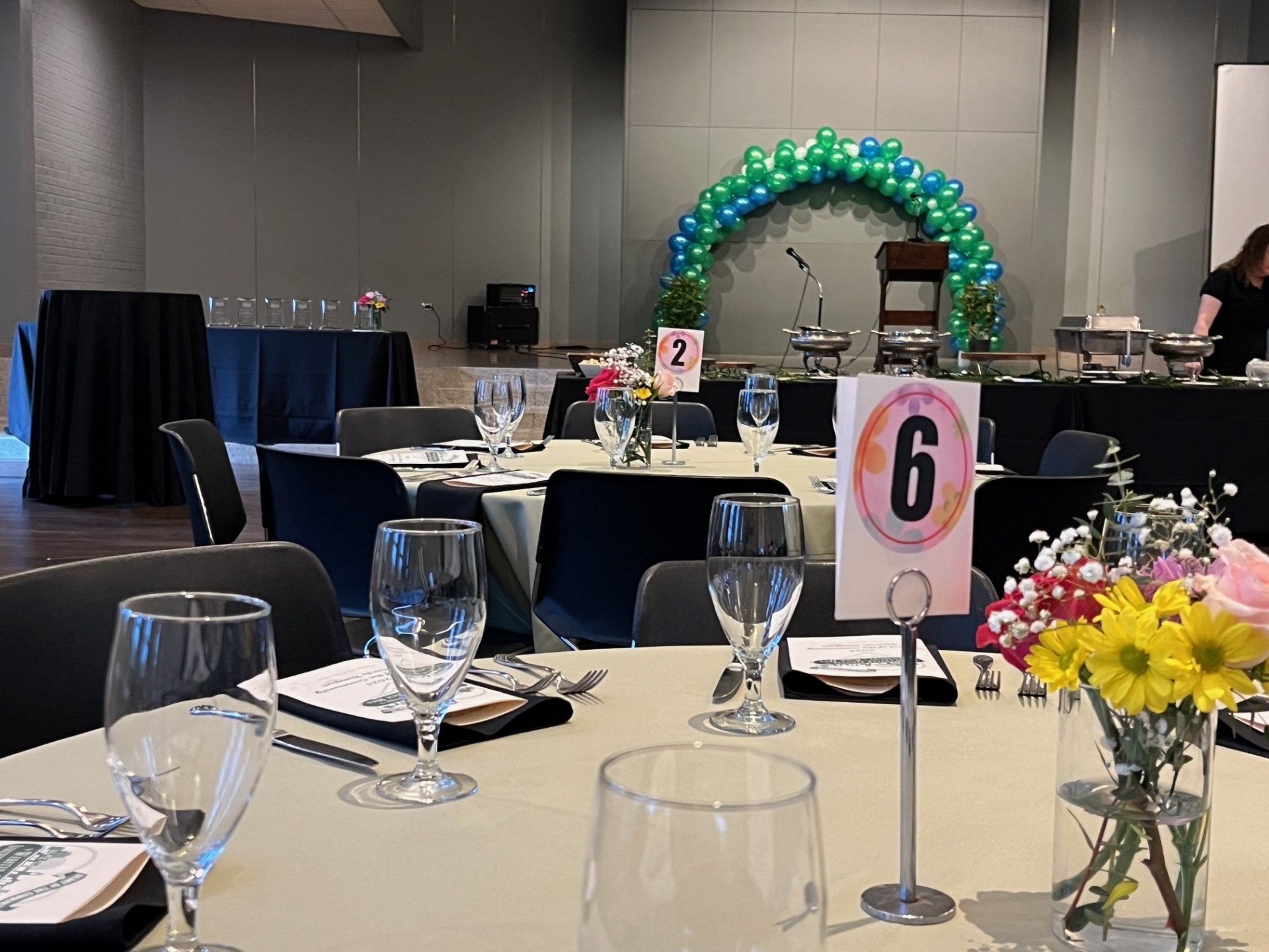 Event hall set up with round tables, floral centerpieces, and a stage with a balloon arch.