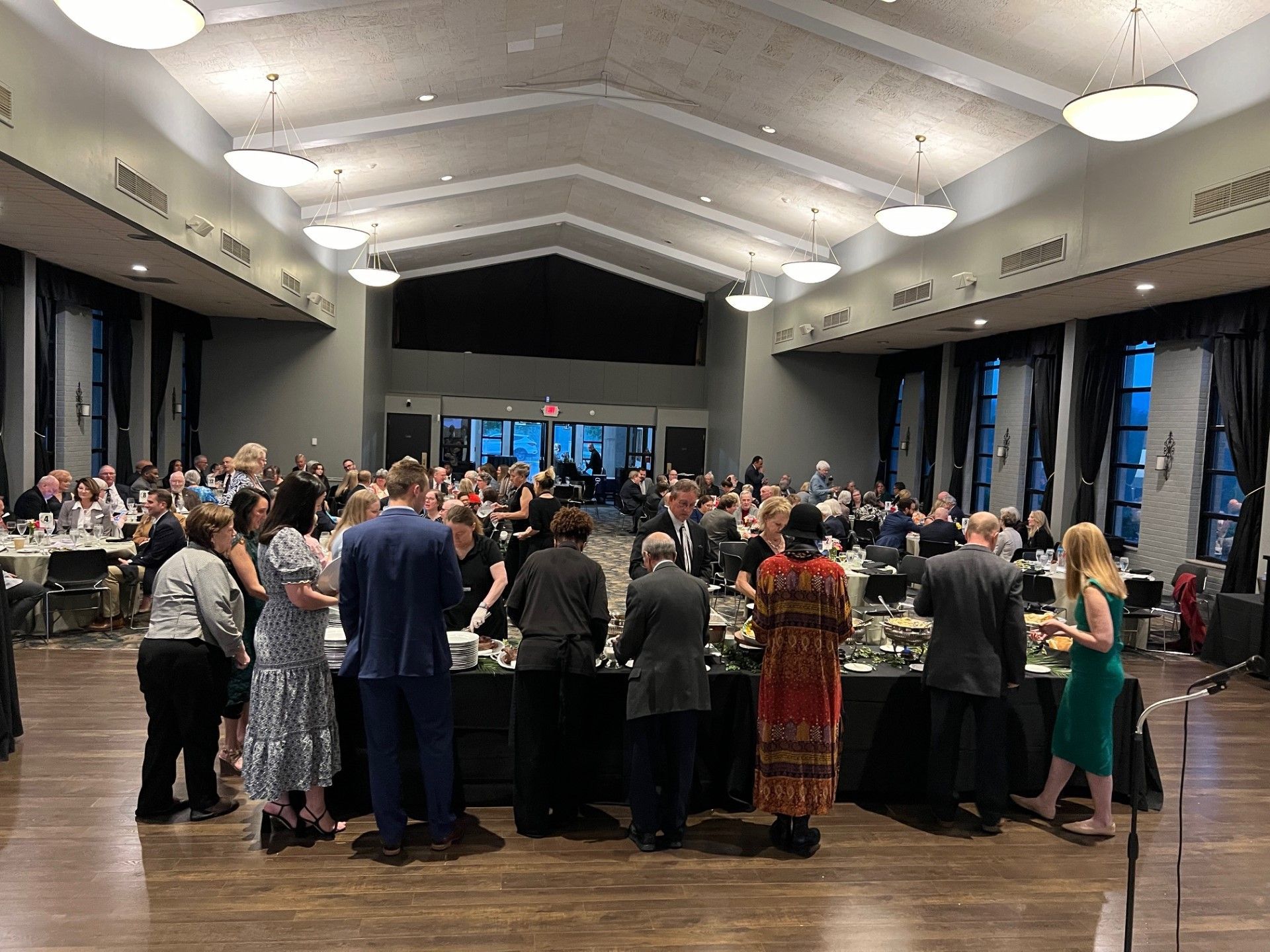 Large event hall, people gathered near buffet tables and seated at tables. Dim lighting, formal attire.