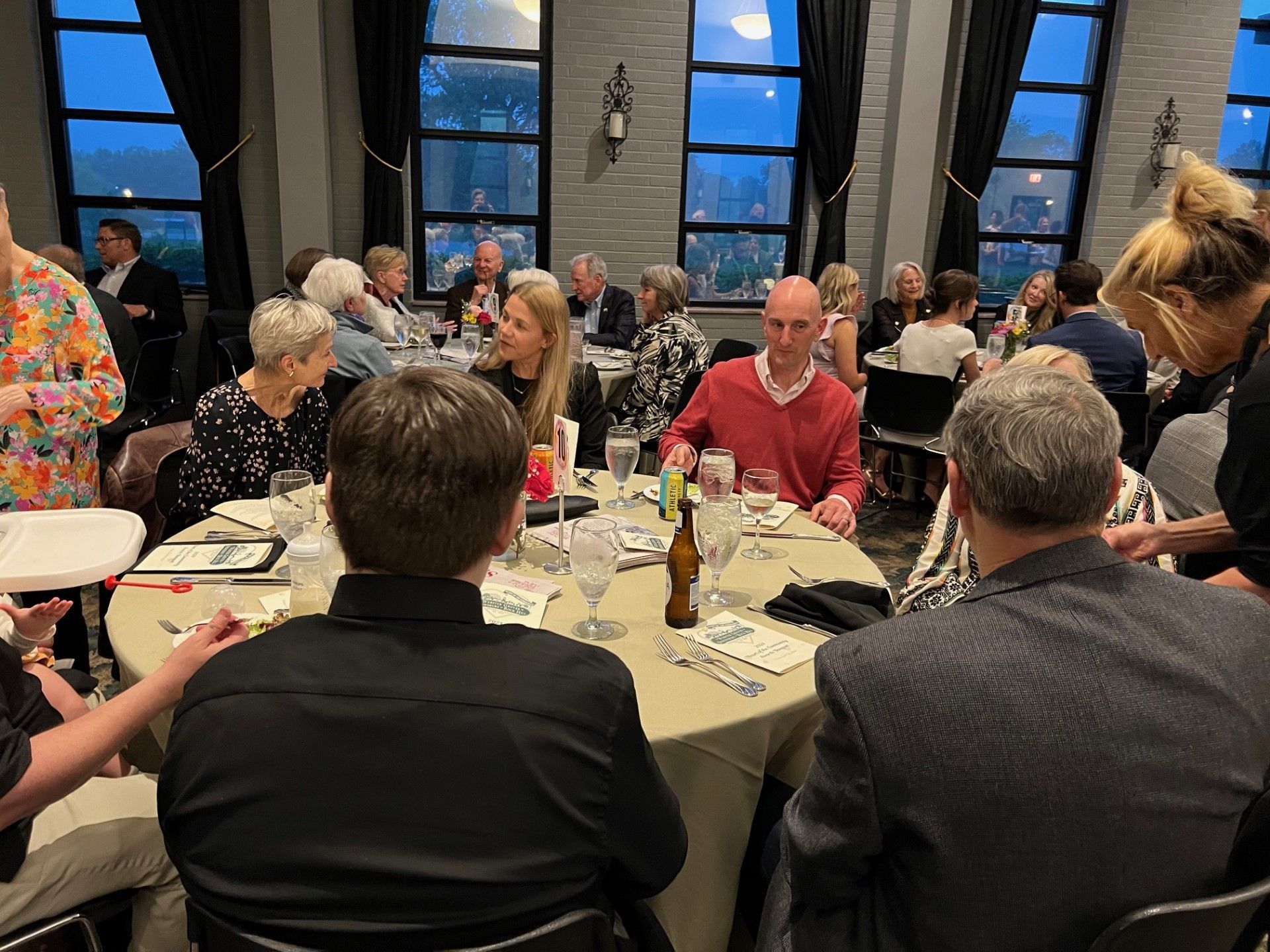 People seated around a large table in a restaurant. Sunlight streams through the windows in the background.