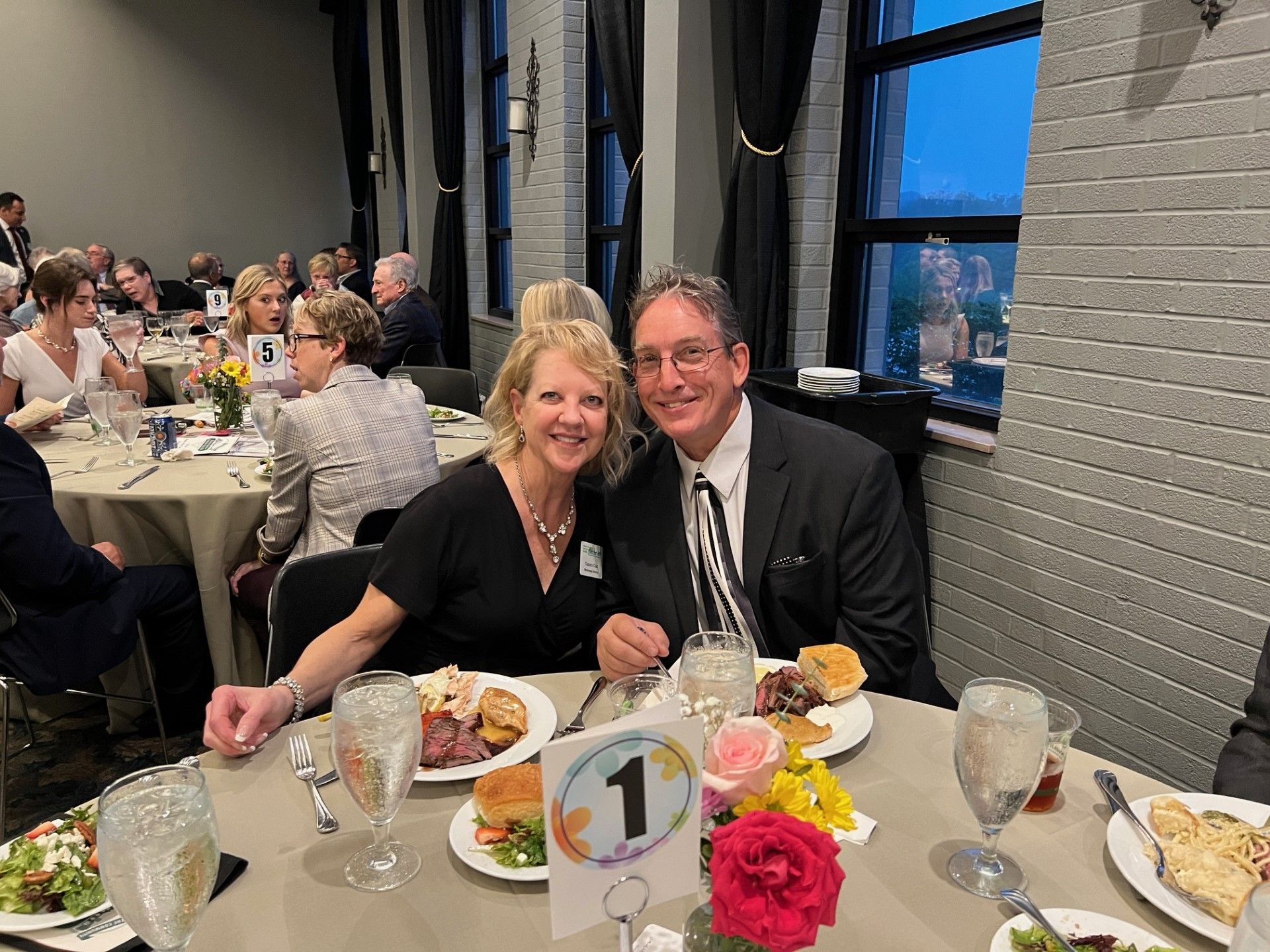Couple smiles at table in banquet hall. They have plates of food, flower centerpiece.