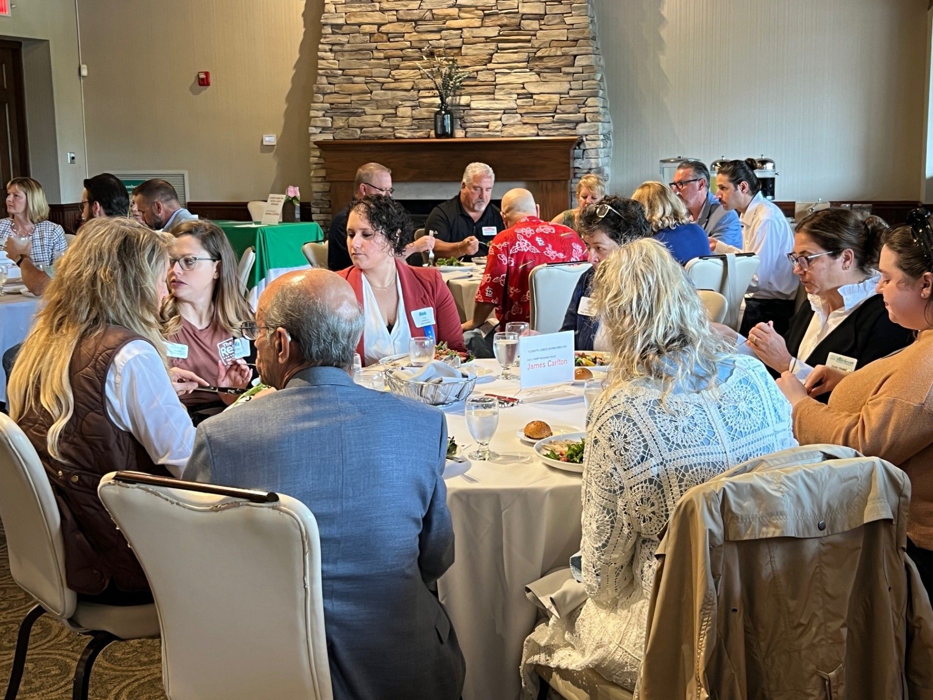 People seated at round tables in a restaurant, eating and conversing. Interior with stone wall and fireplace.