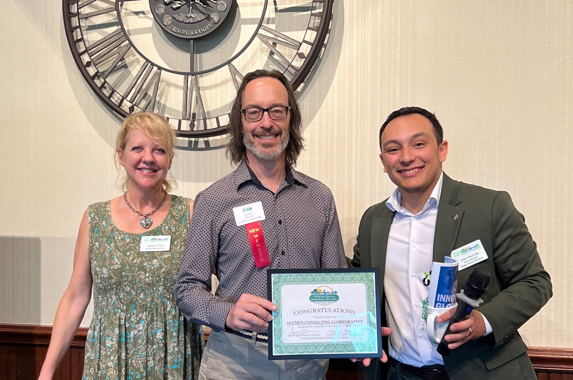 Three people holding an award in front of a clock on a wall.