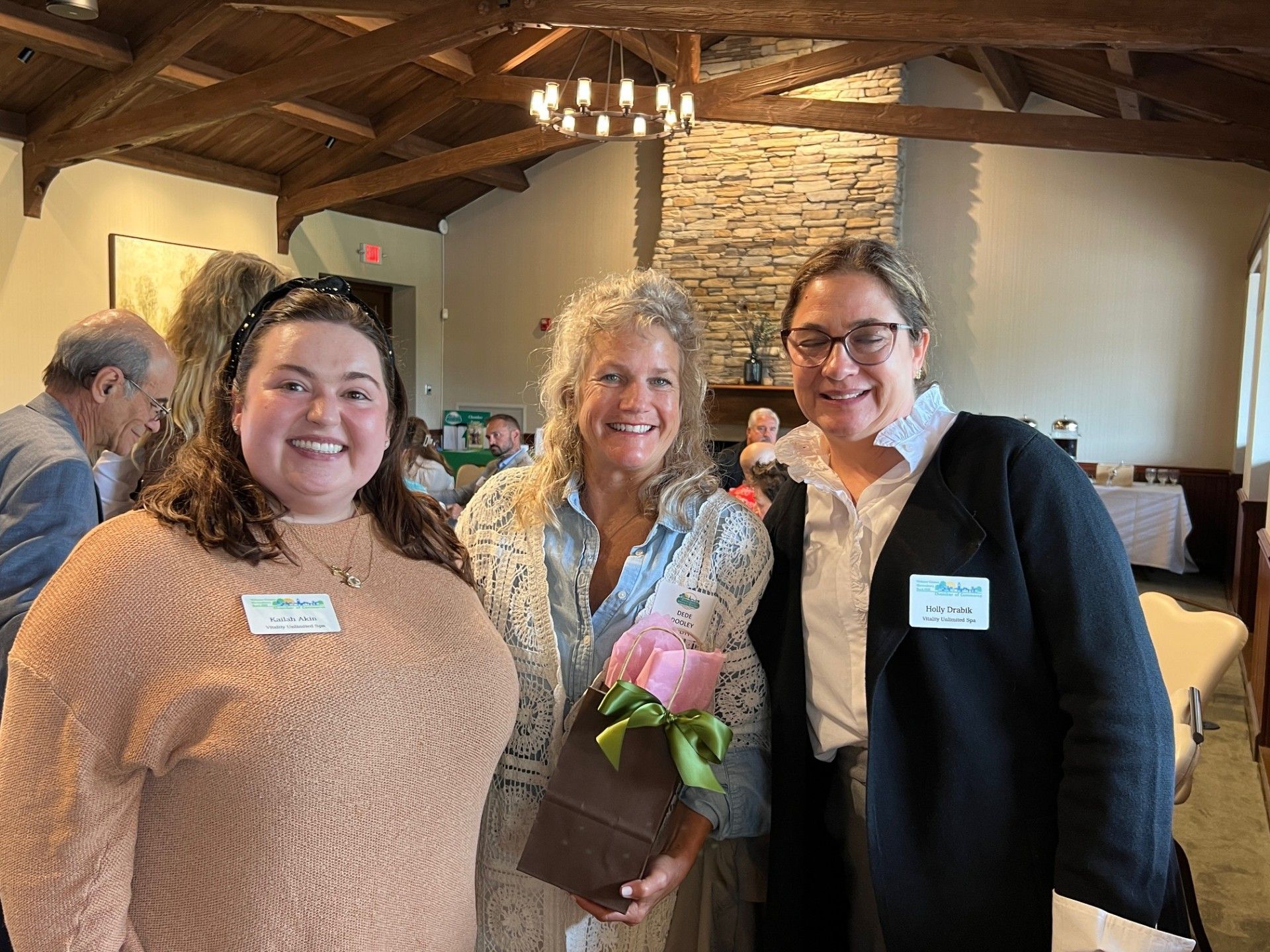 Three women smiling, holding gift and flowers in a room with a light fixture and stone fireplace.