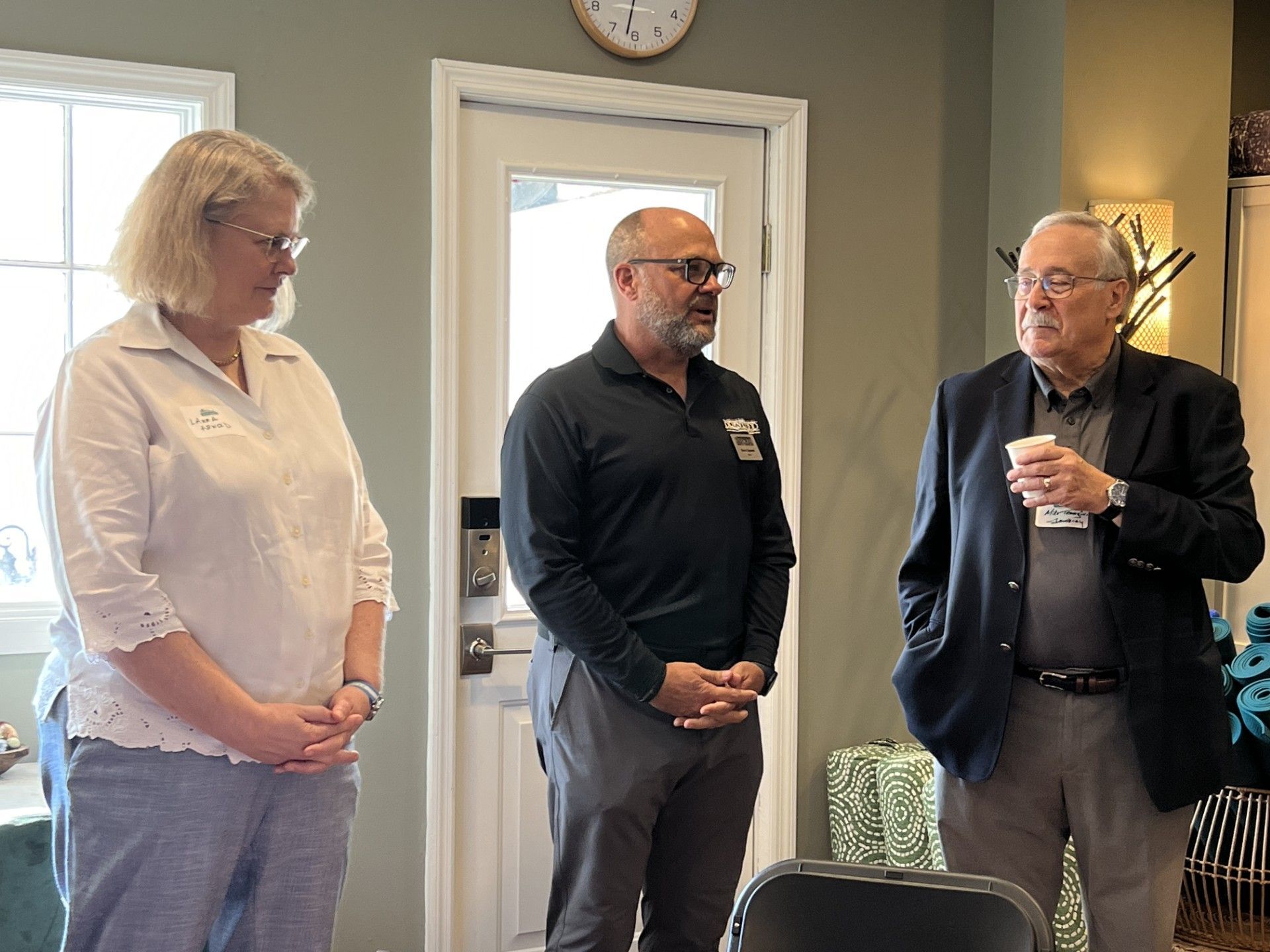 Three people in a room, two men and one woman, talking. One man holds a cup.