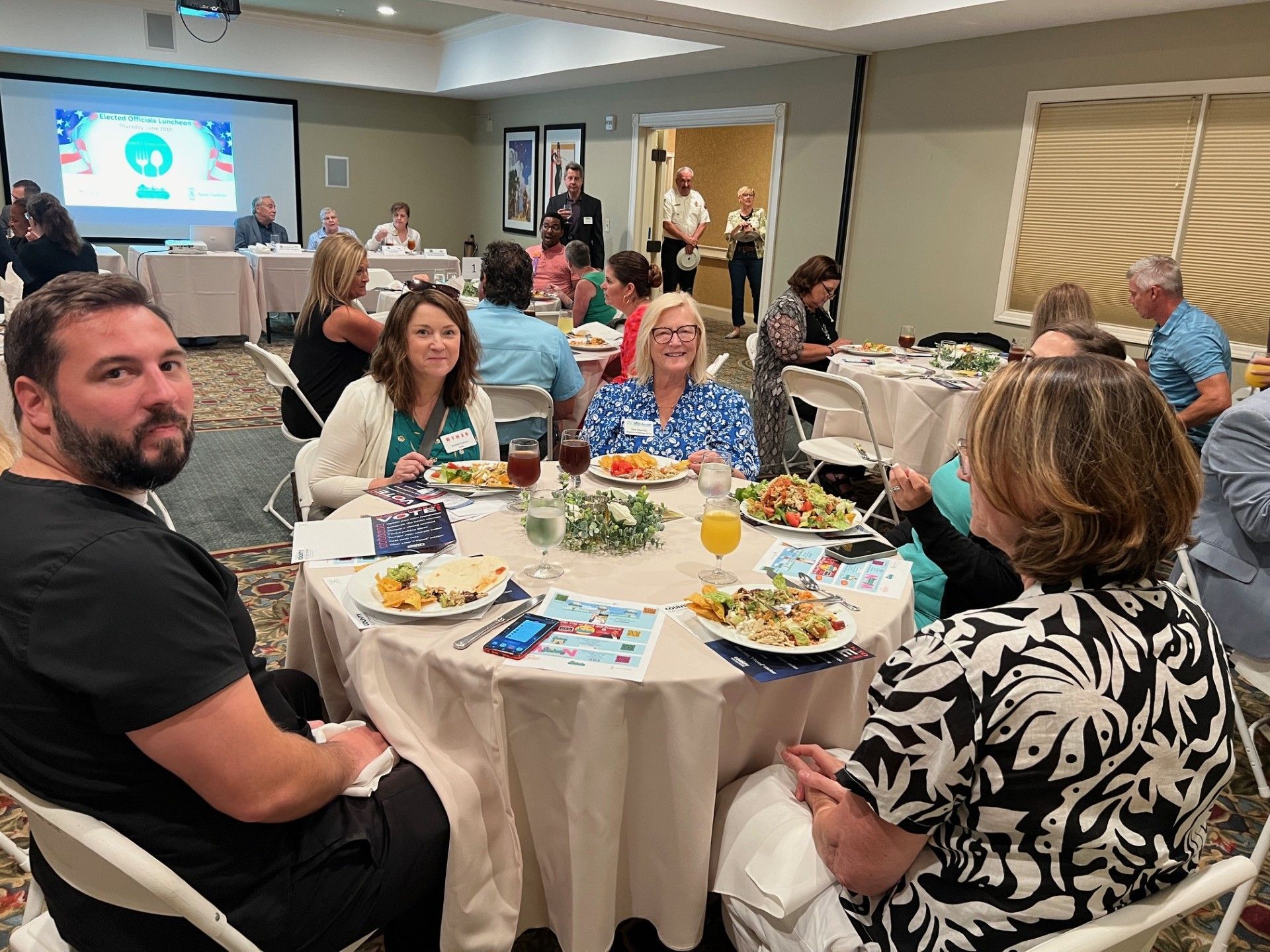 People seated around tables at a catered event, some eating. A presentation screen is in the background.