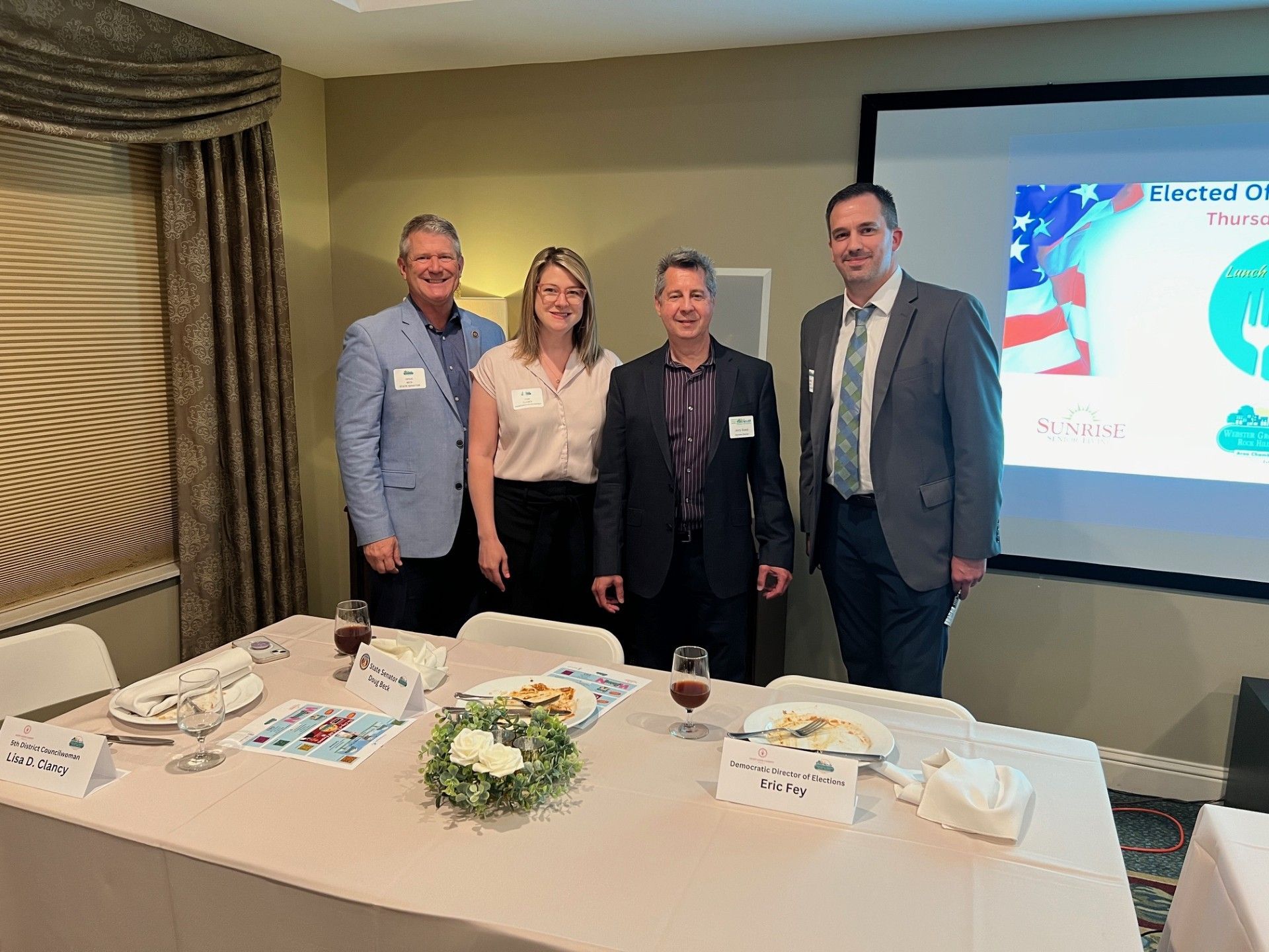 Four people stand behind a table with food and wine. A projector screen displays a logo.