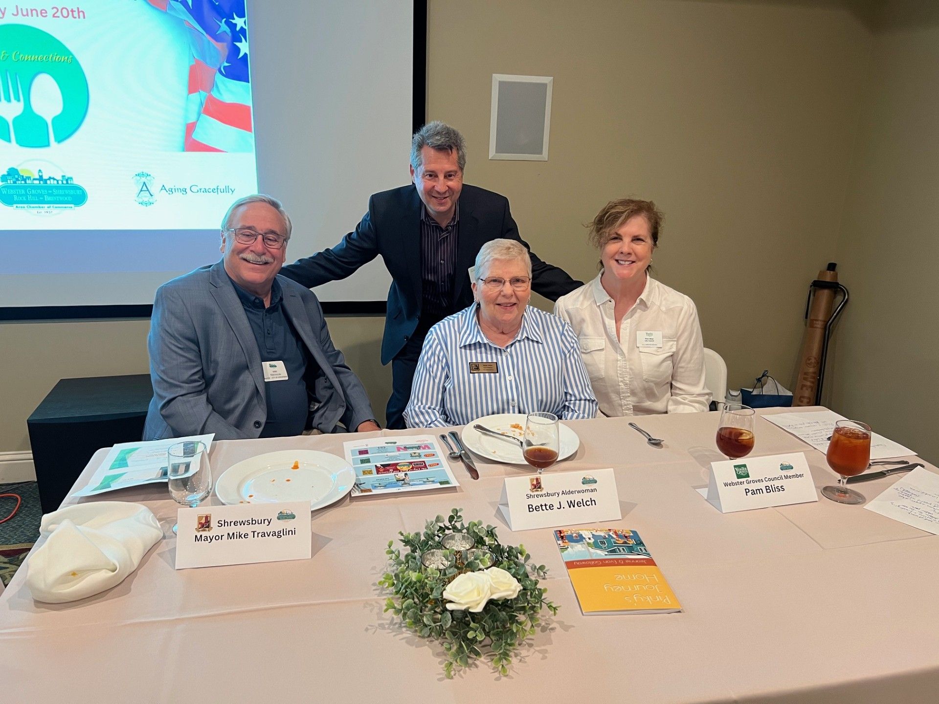 Four people at a table, two seated, two standing behind. A banner is in the background.