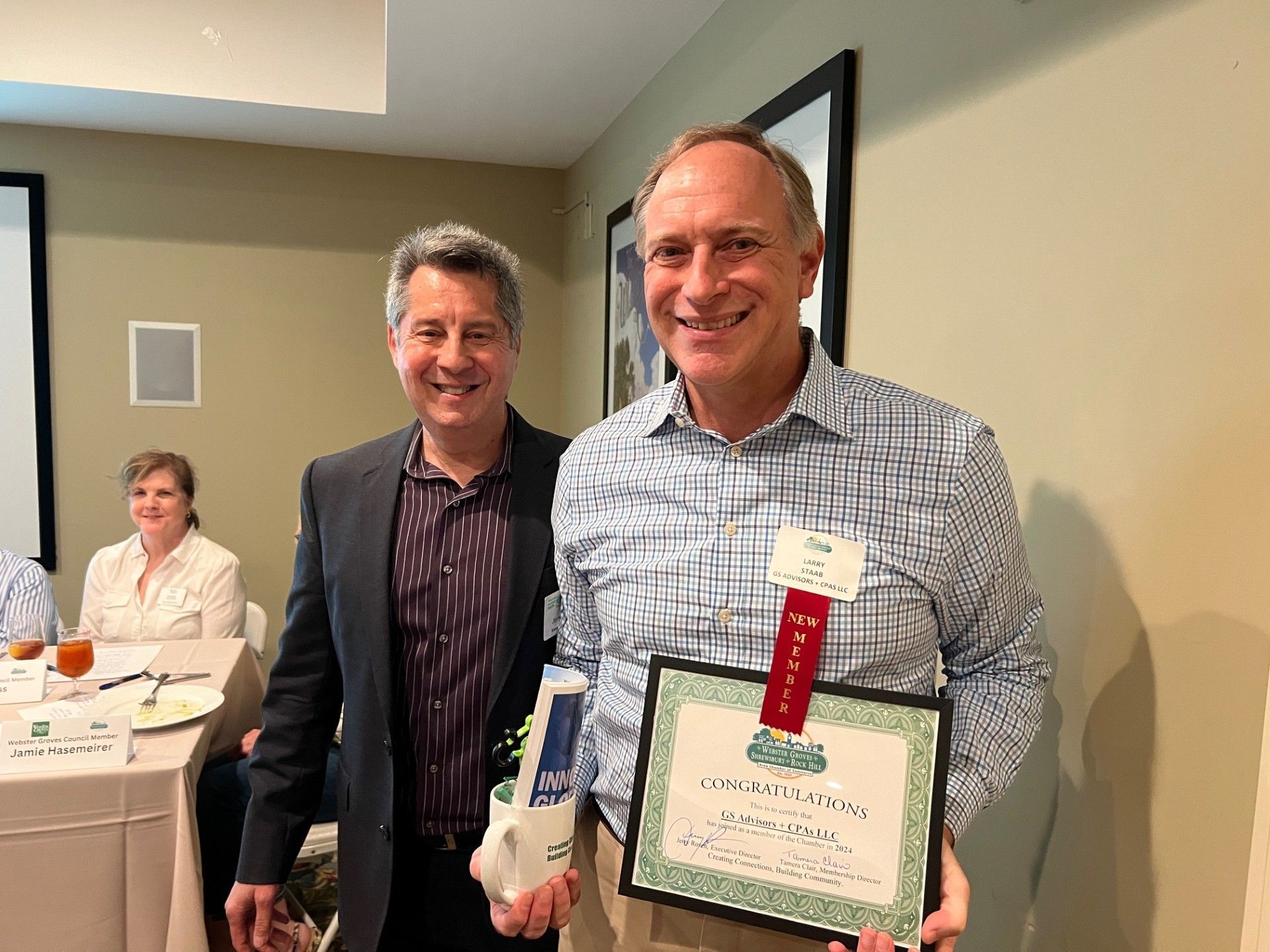 Two men smiling, one holding an award with a red ribbon. They are inside a room with others seated at a table.