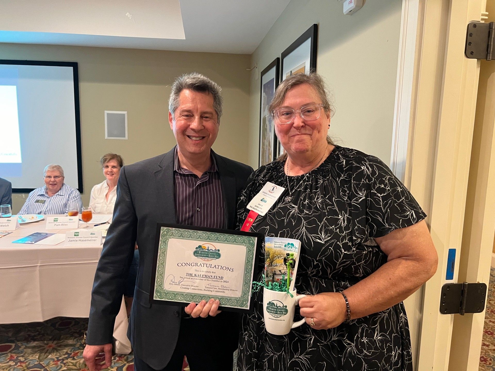 Man in suit presents award to woman holding gift basket and mug. Conference room setting.
