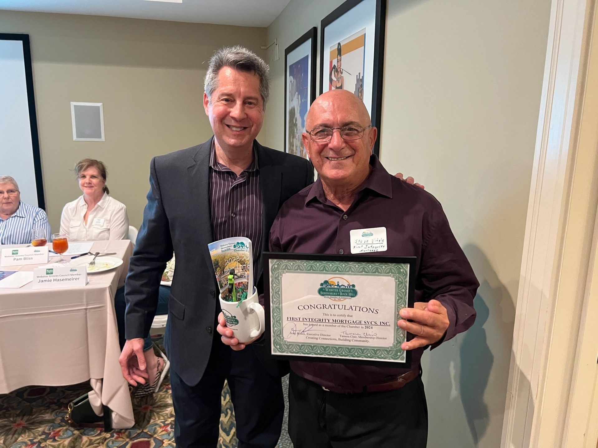 Two men in a room, one holding a certificate and the other holding a gift bag. They are smiling.