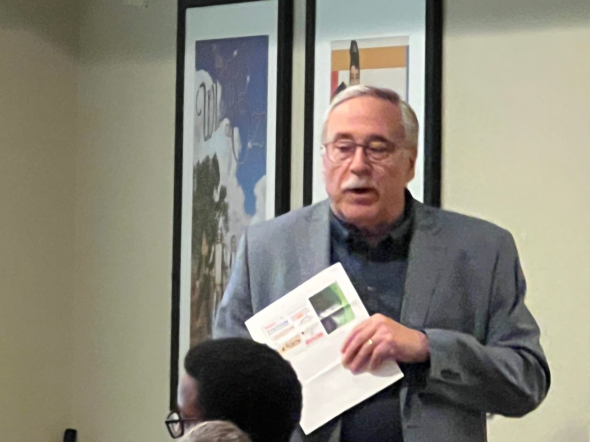 Man in gray jacket speaks, holding papers. Two framed posters behind him.