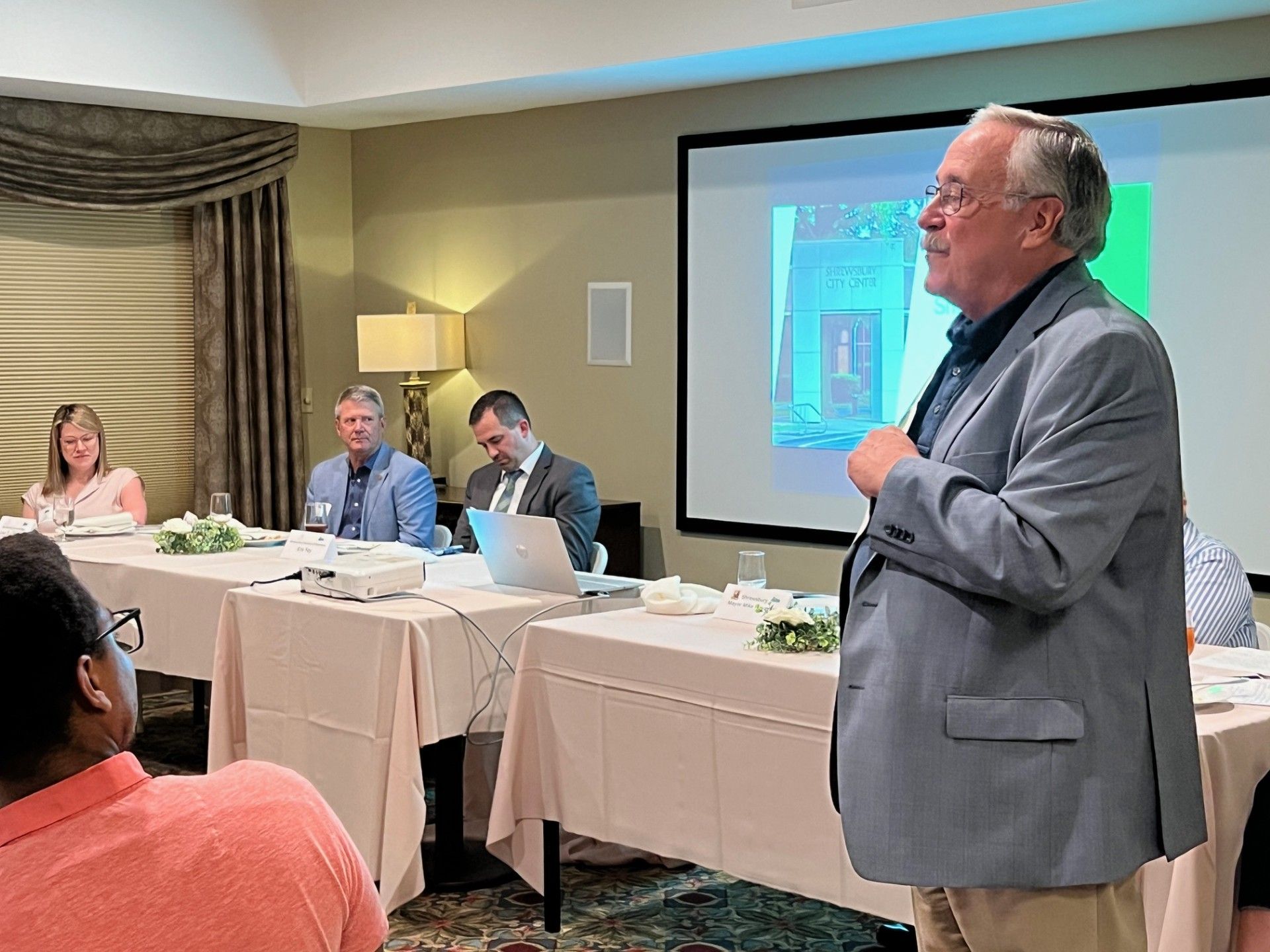 Man speaking at a conference. Others sit at a table. Presentation screen visible.