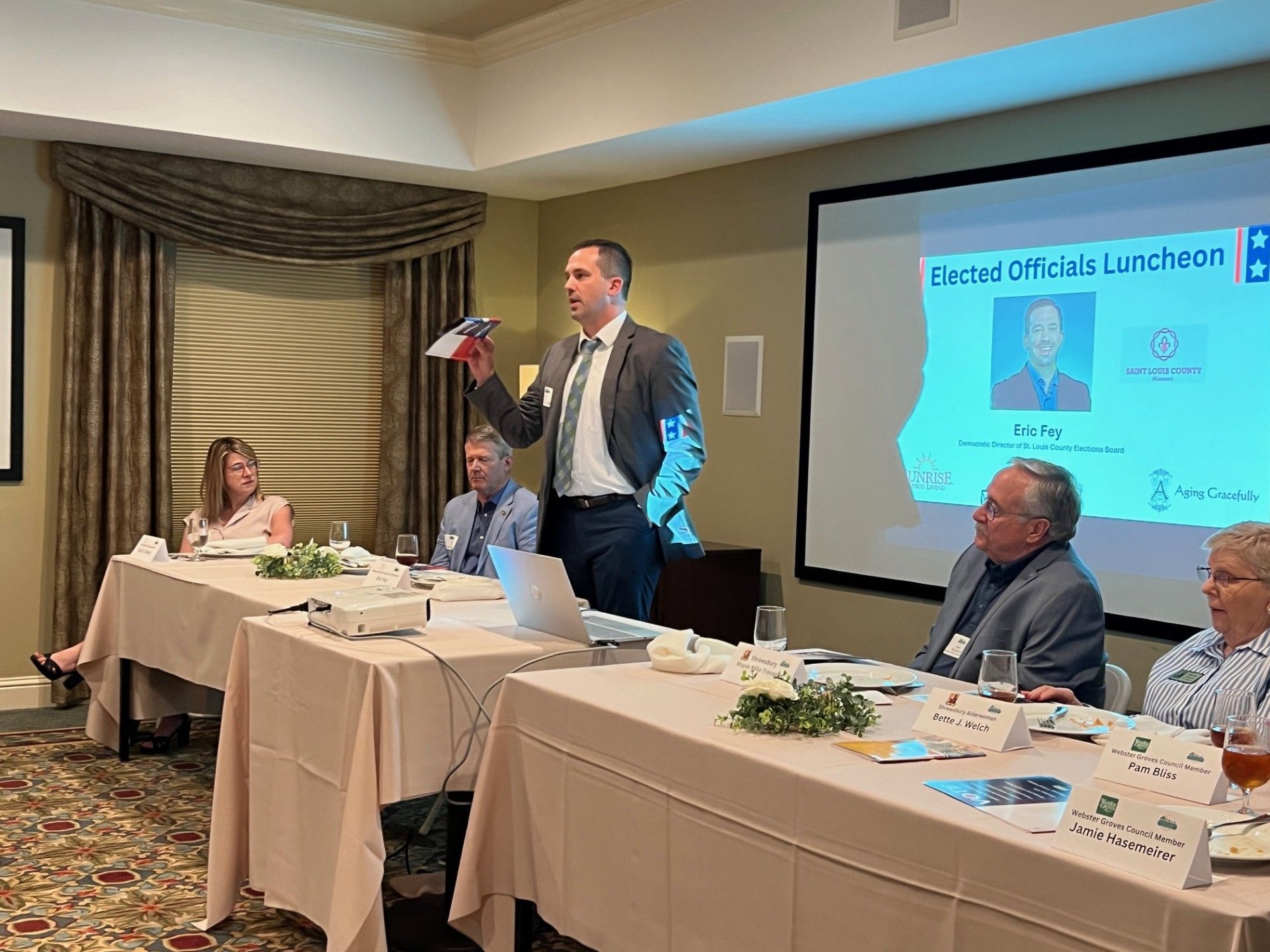 Man speaking at a luncheon, holding an object. Others sit at a table in front of a projection screen.