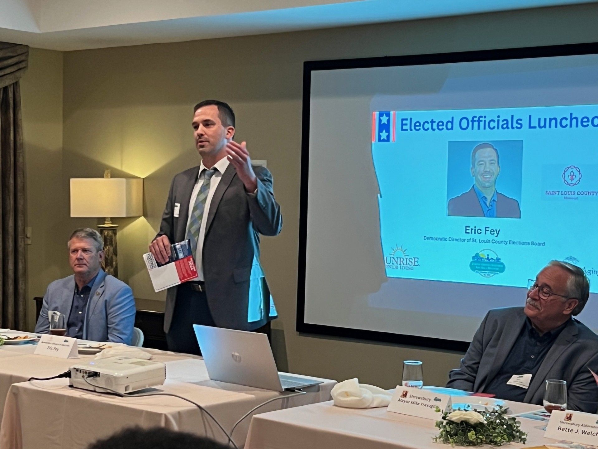Man speaking at a luncheon. Two men sit at a table. Presentation screen in the background.