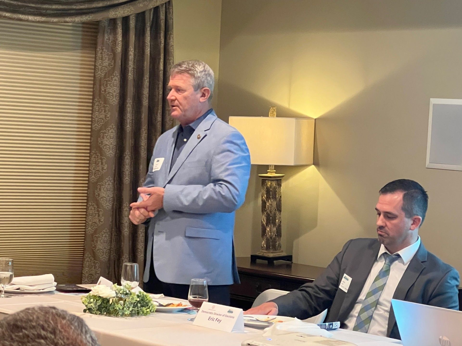 Man in a light blue jacket speaks at a table, another man sits beside him; indoor, daytime.