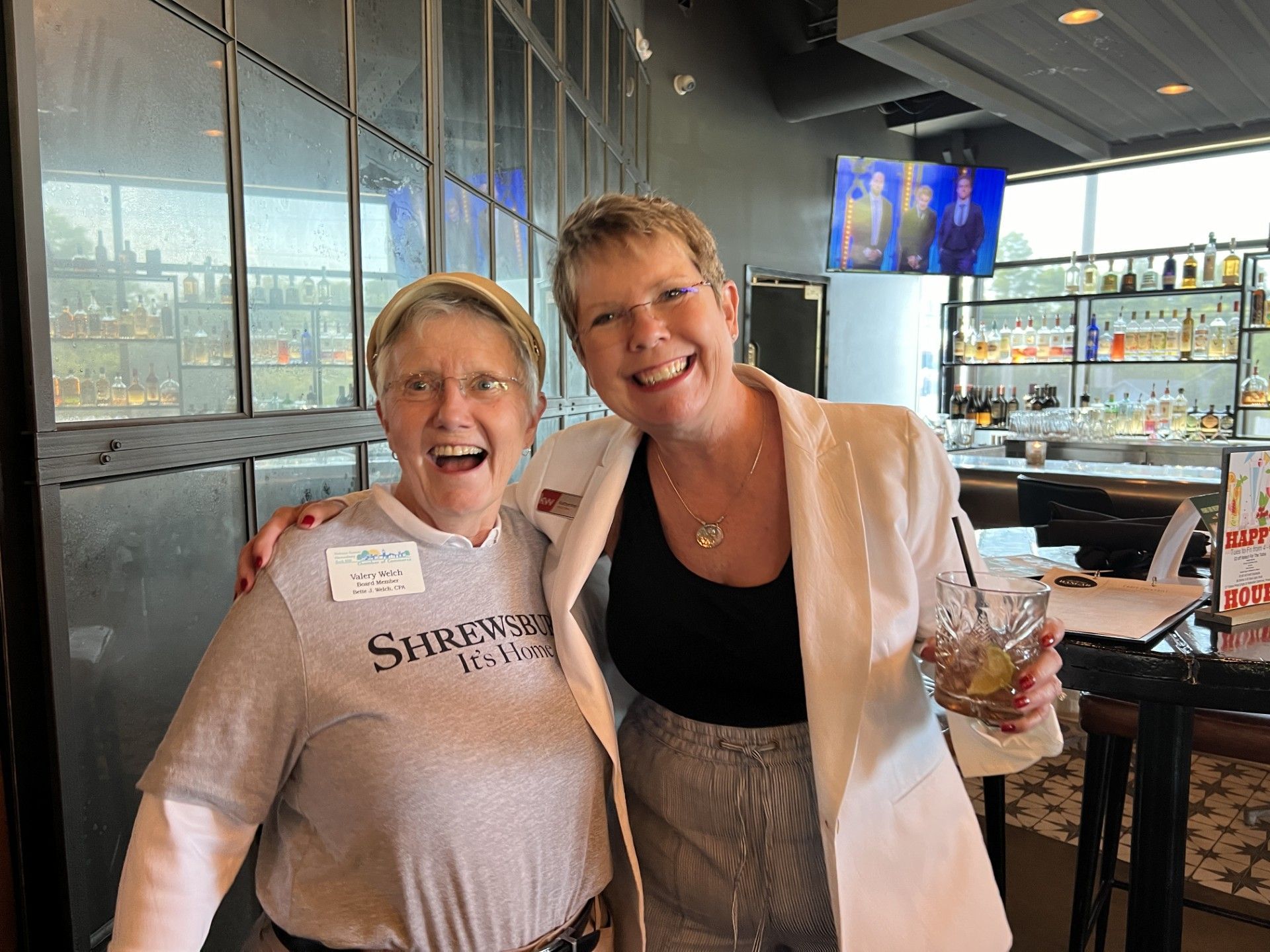 Two women smiling, posing at a bar. One holds a drink, the other wears a name tag.