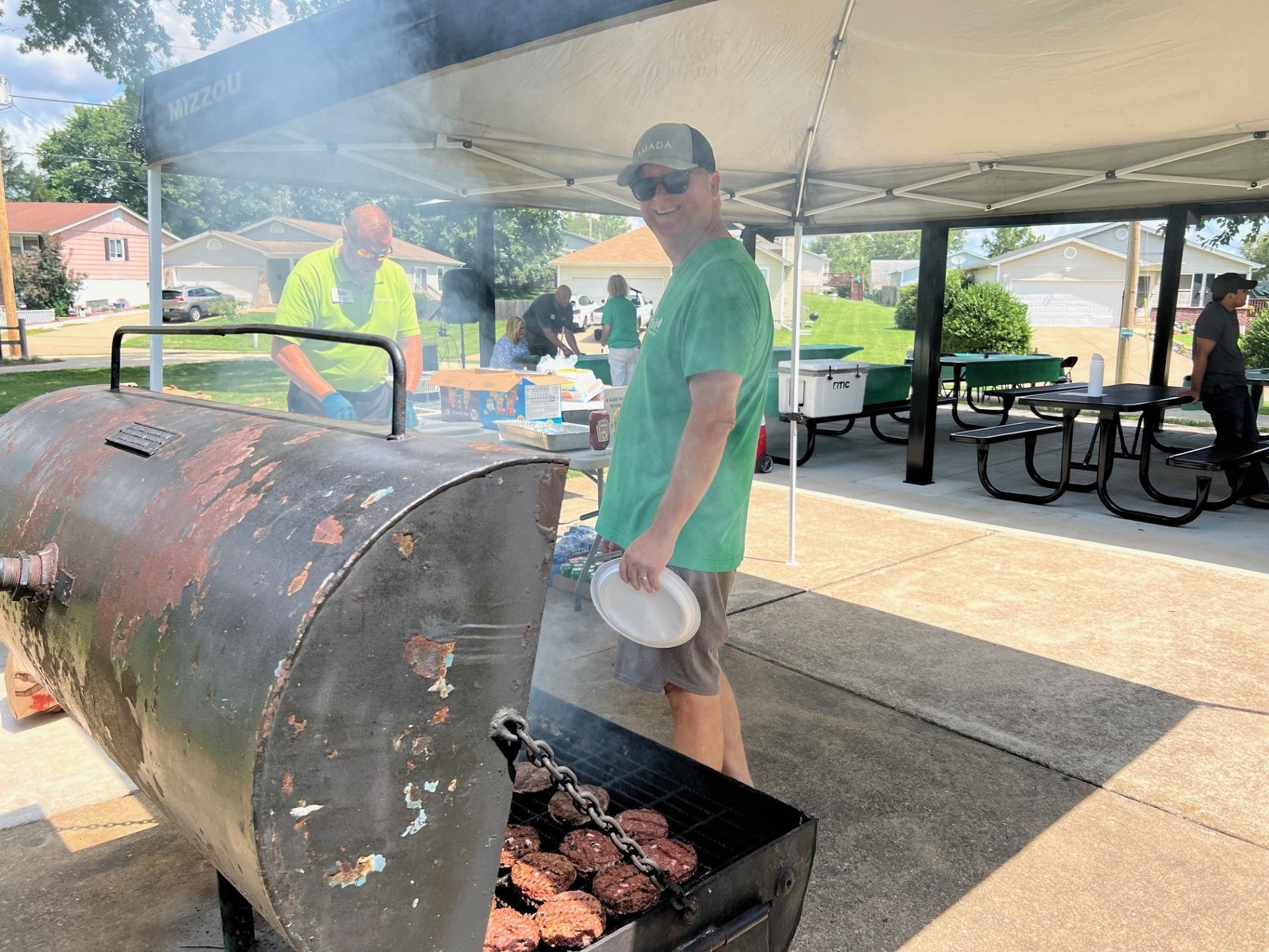 Man grilling burgers at a cookout, holding a plate. Others are nearby under a shelter.