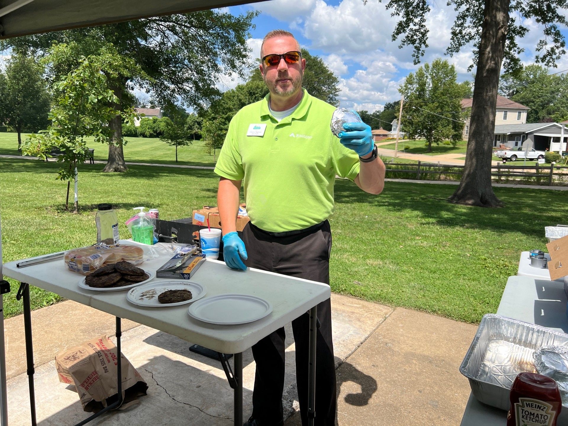 Man in green shirt, sunglasses, and gloves holds food over a table with food on it outdoors.