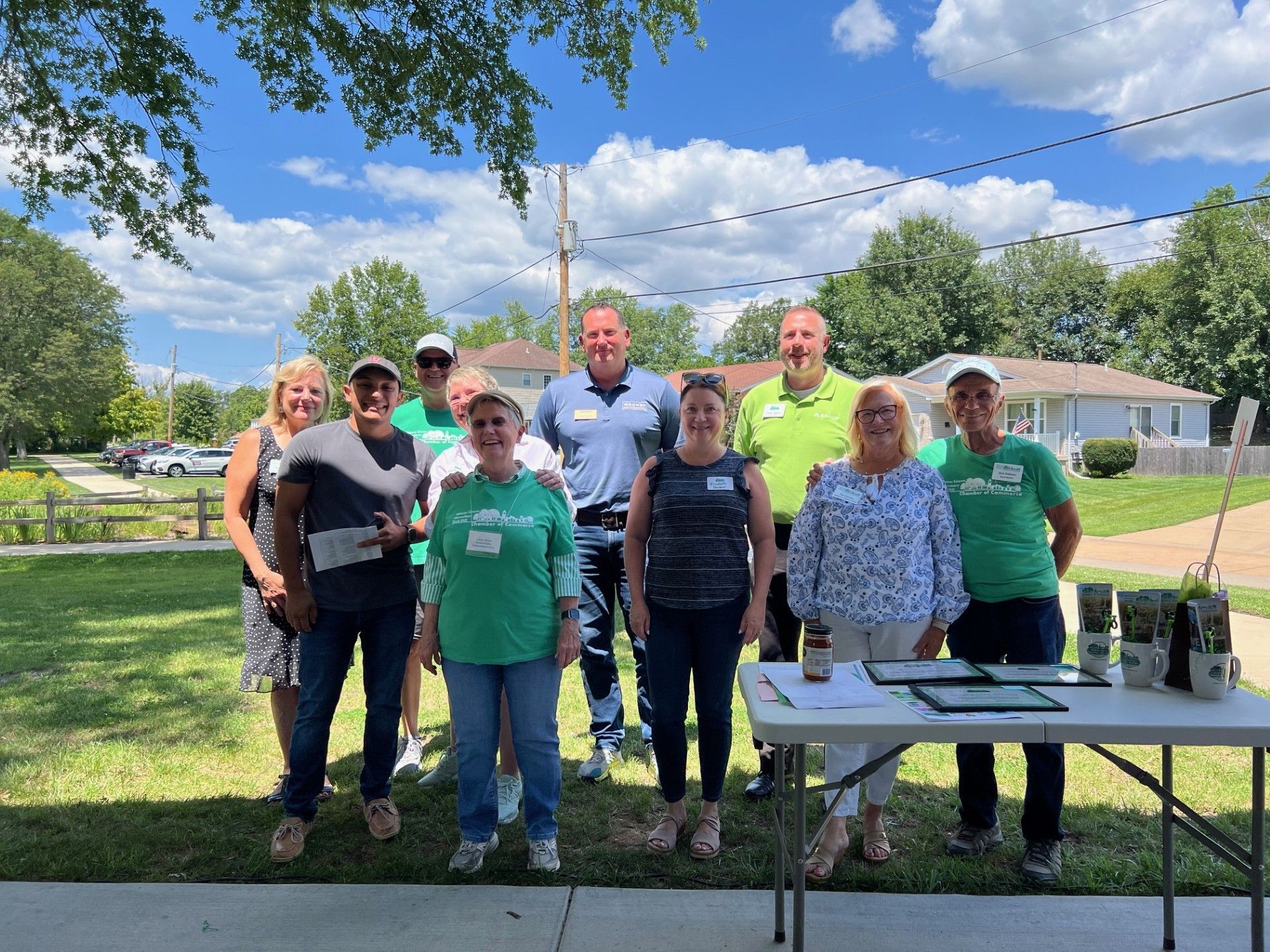 Group of people smiling outdoors, standing near a table with materials under a sunny sky.
