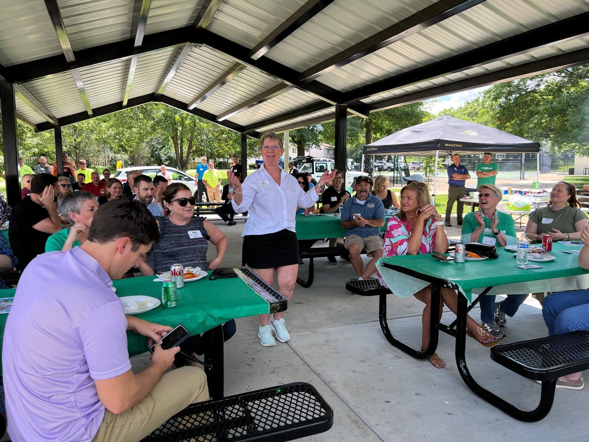 People gathered at outdoor picnic tables under a covered structure, listening to a woman speaking.