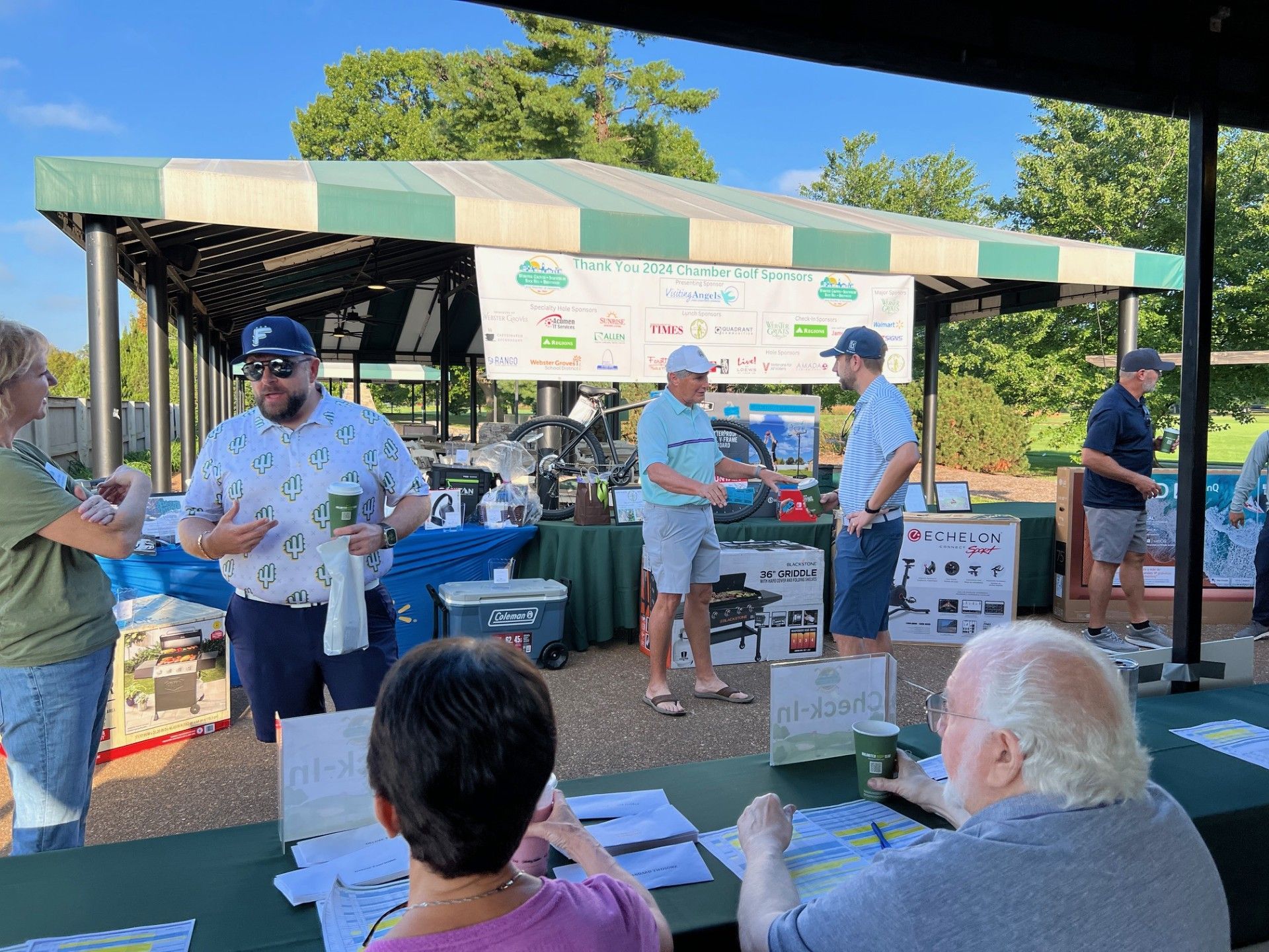 People at an outdoor golf event registration table. Merchandise and signs are visible.