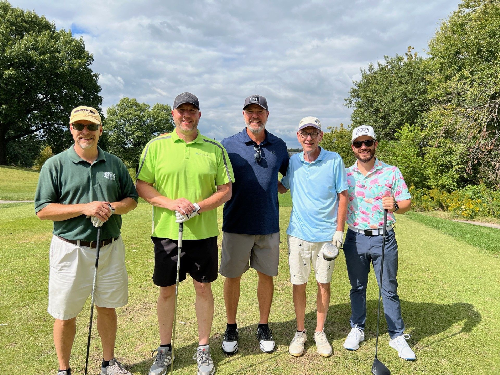 Five men on a golf course posing for a photo. They hold golf clubs, wearing shorts, and shirts.