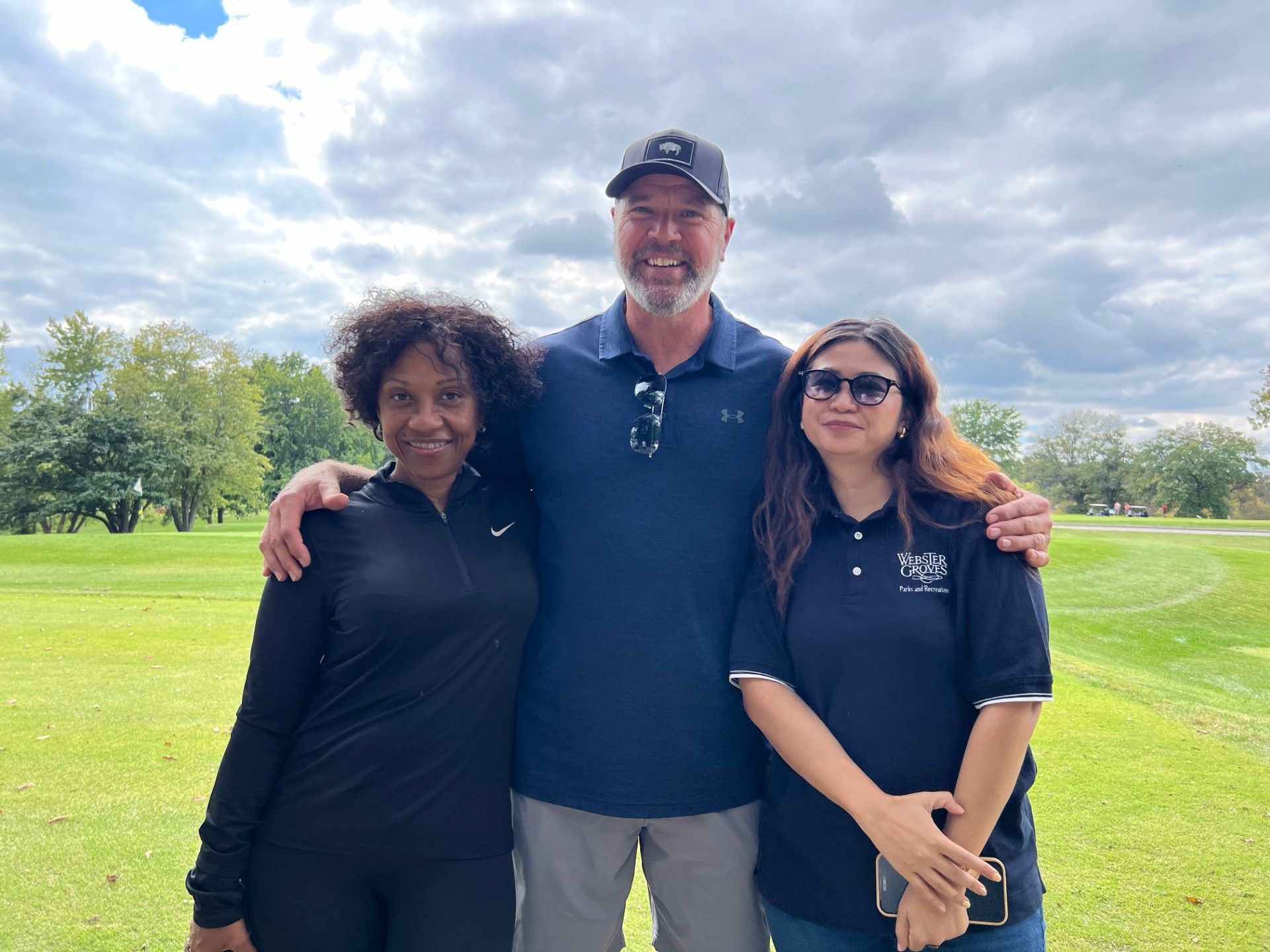 Three people pose on a golf course. Man in a hat has his arms around two women. Green grass and trees in the background.