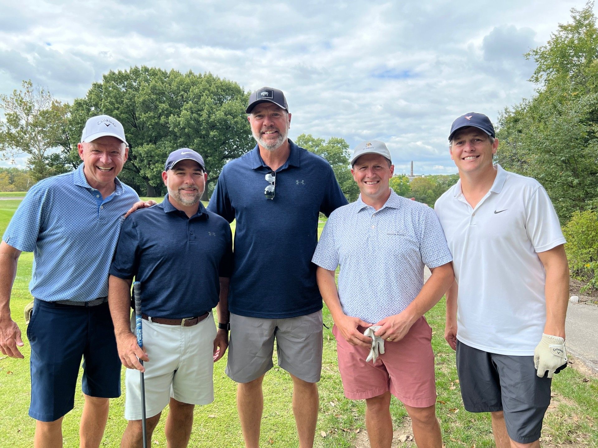 Five men in golf attire pose on a golf course, smiling at the camera.