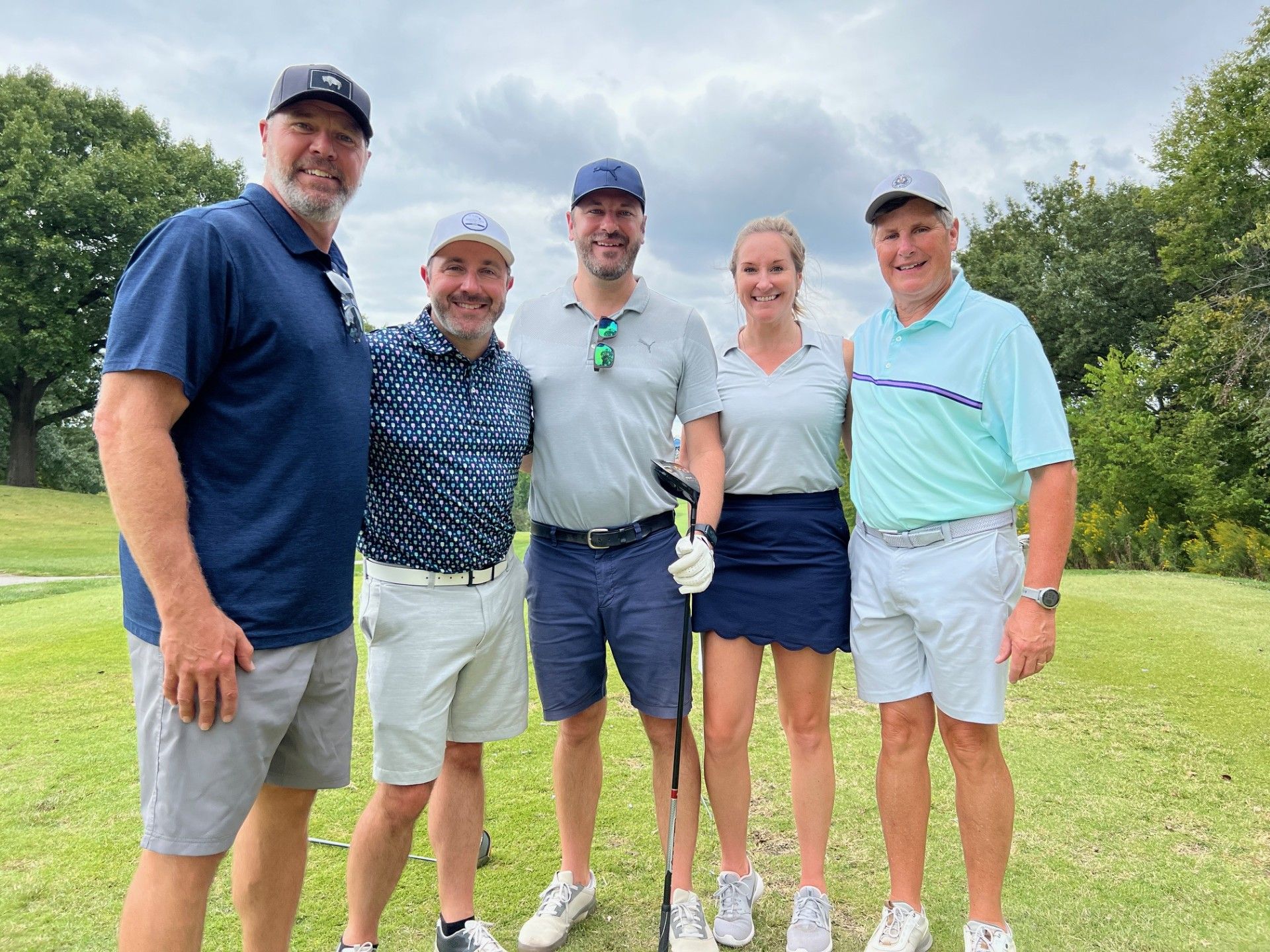 Five people smiling on a golf course. They are wearing golf attire, holding clubs. Cloudy sky.