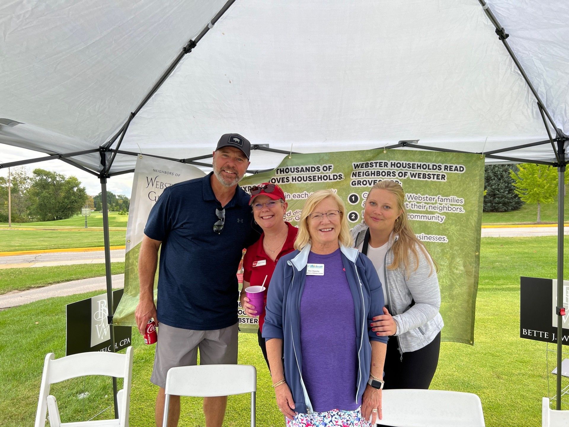 Four people smiling under a canopy at an outdoor event, with a promotional banner in the background.