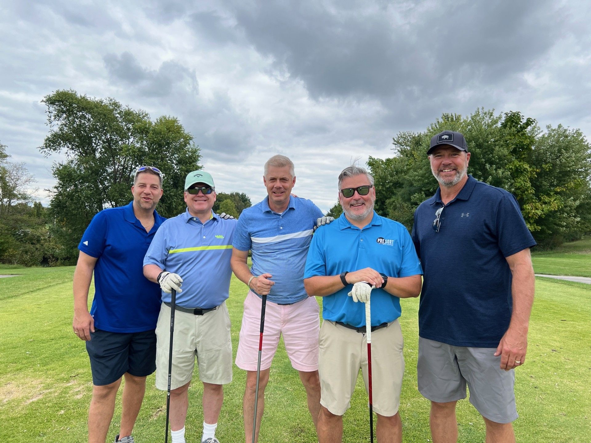 Five men standing on a golf course, holding clubs. Cloudy sky, green grass, and trees are in the background.