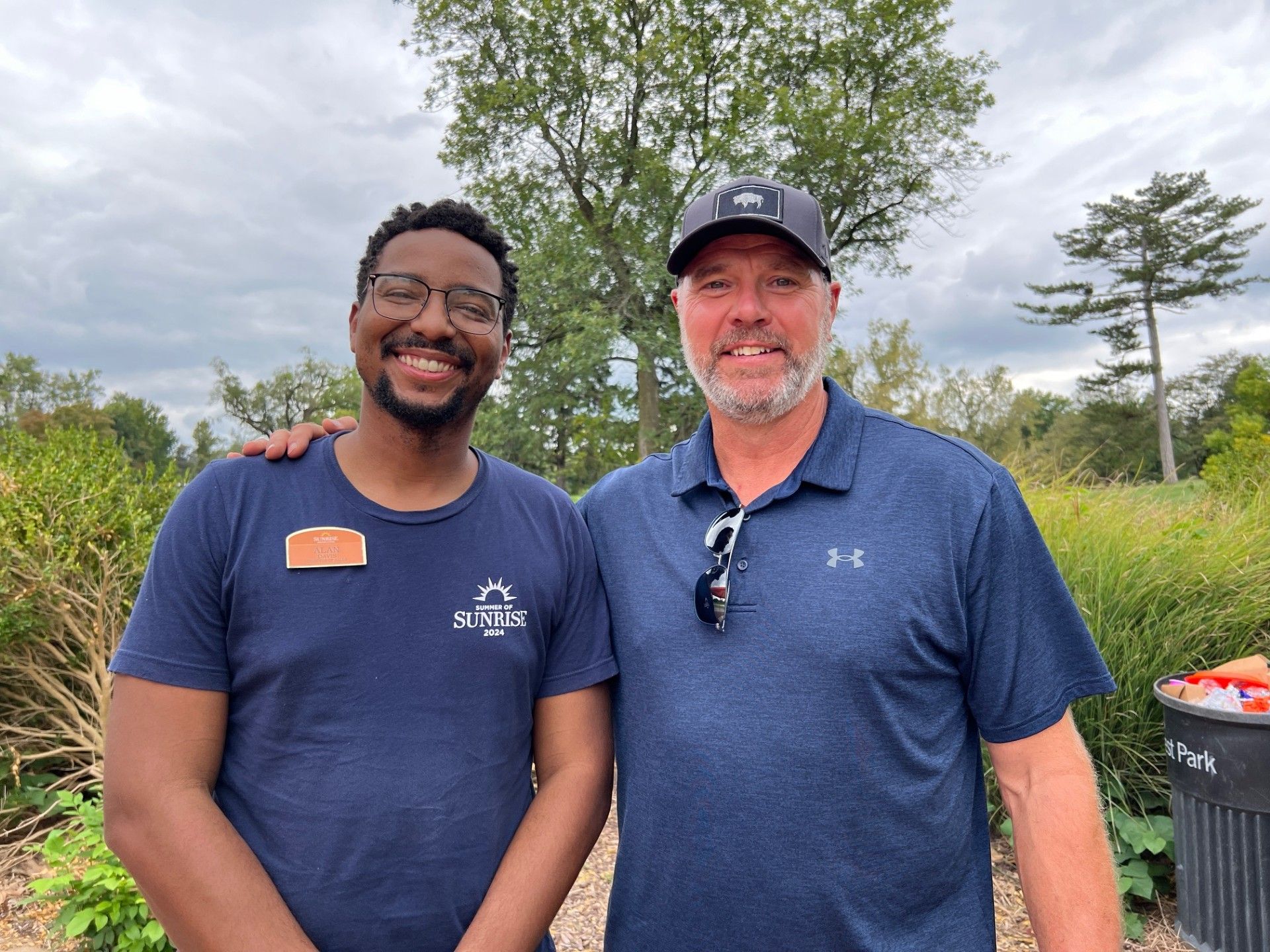 Two men smiling outdoors; one has arm around the other's shoulder. Green foliage and overcast sky in background.