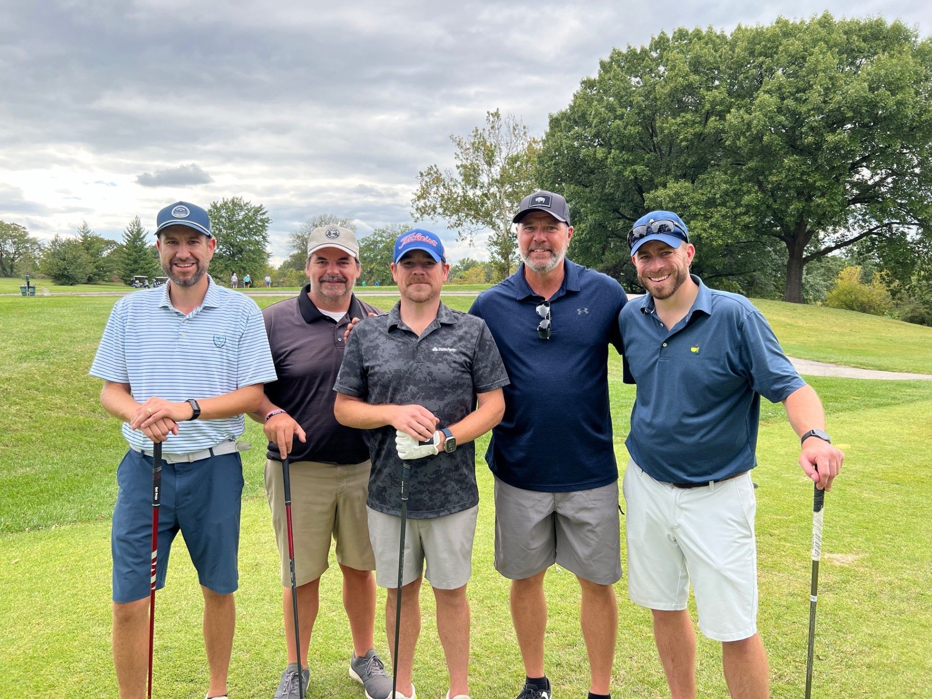 Five men smiling, posing on a golf course, holding clubs. Cloudy sky, green grass, and trees in the background.
