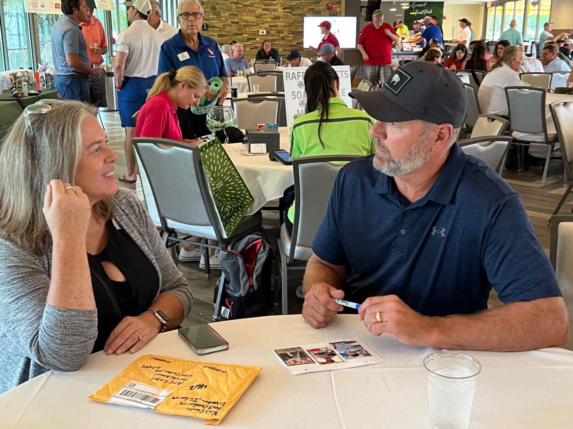 Two people at a table talking. Man signs something. Tables and people in a large room.