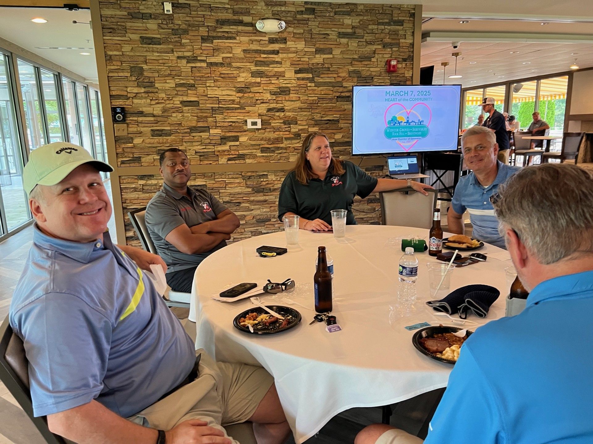 Group of people seated around a table, some smiling, with food and drinks. A screen displays text.