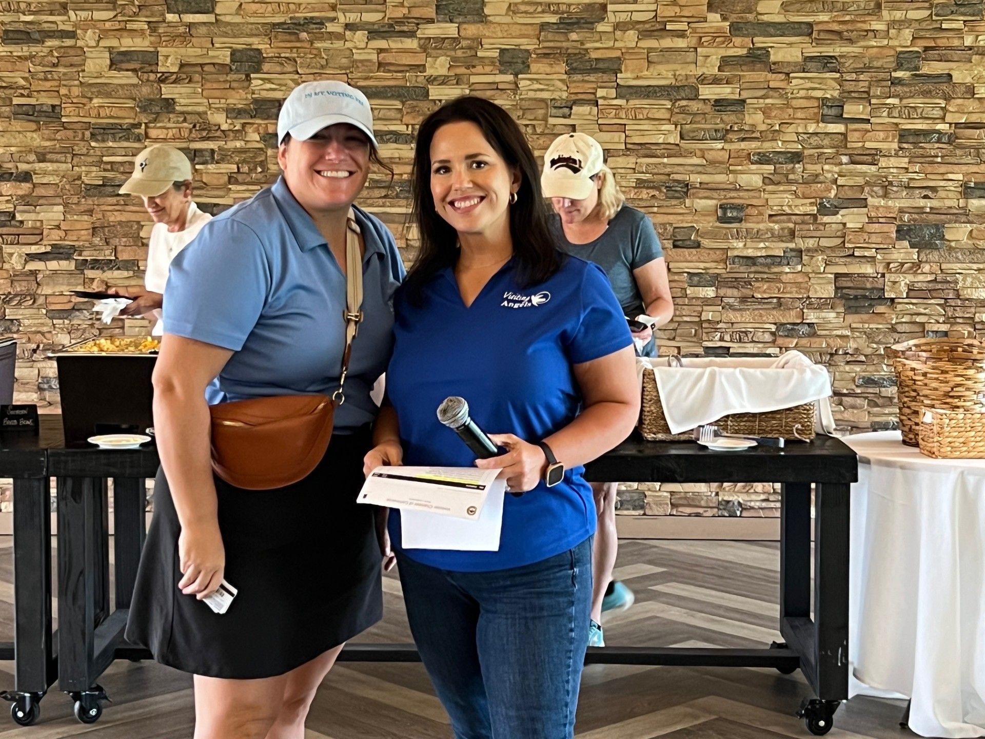 Two women smiling, posing at an outdoor event with a table and snacks. One holds a microphone and a check.