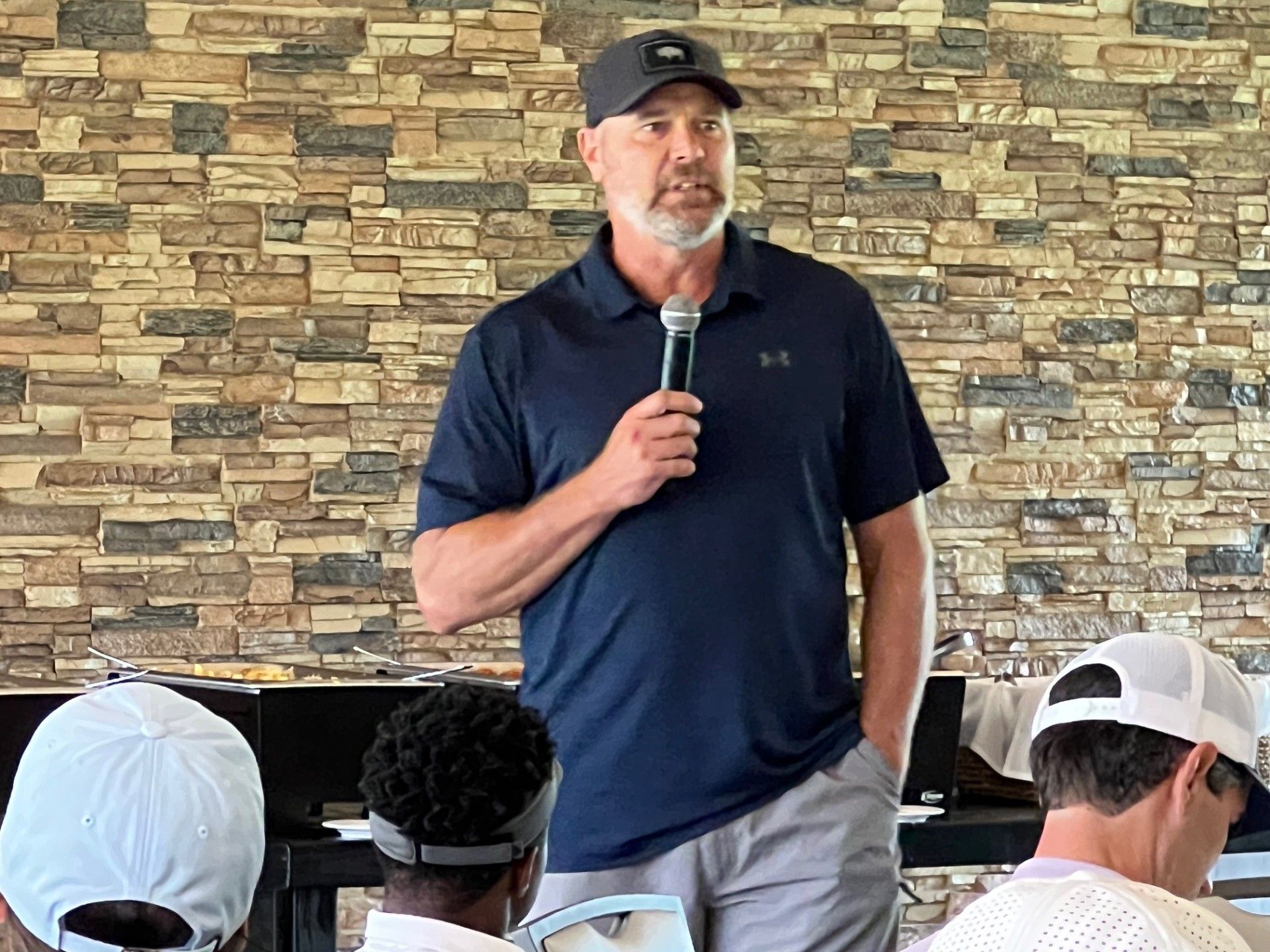 Man in cap, navy shirt, speaking into microphone in front of stone wall.