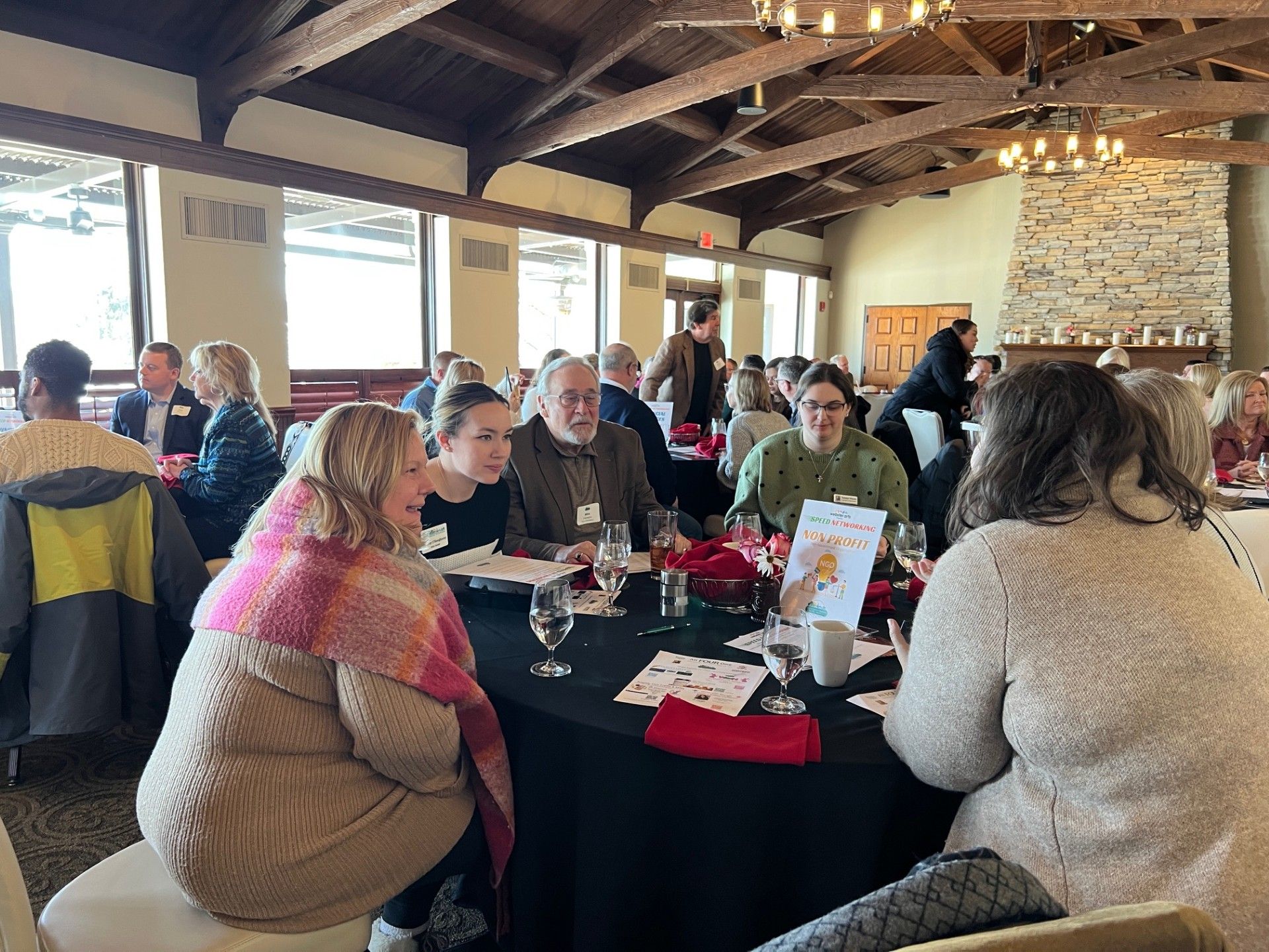People at tables in a brightly lit banquet hall. Some are talking, others look at paper. Red centerpieces.