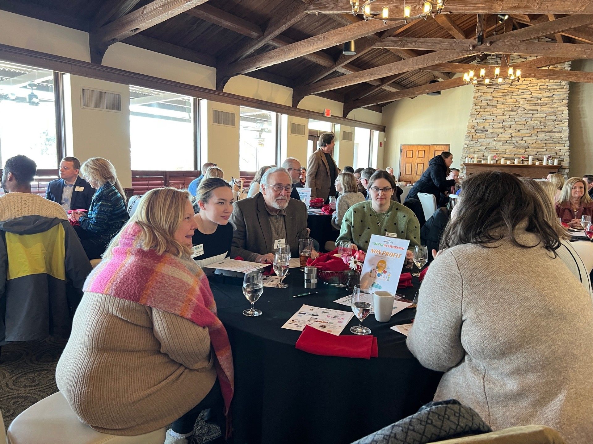 People seated around tables at an event. Some are conversing, others looking at papers. Interior with dark table cloth, fireplace, and exposed beams.