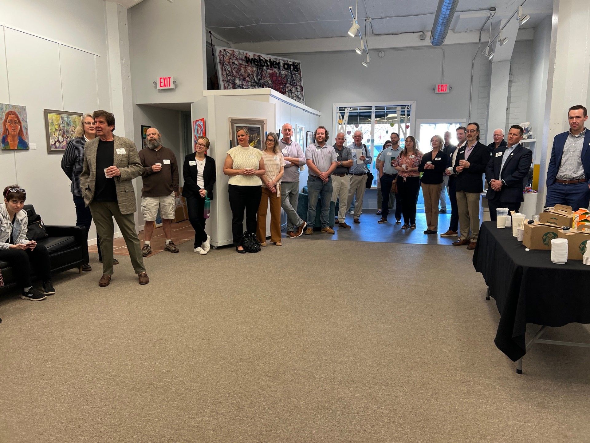 Group of people at an art gallery opening, with someone speaking. Beige carpet, art on the walls.