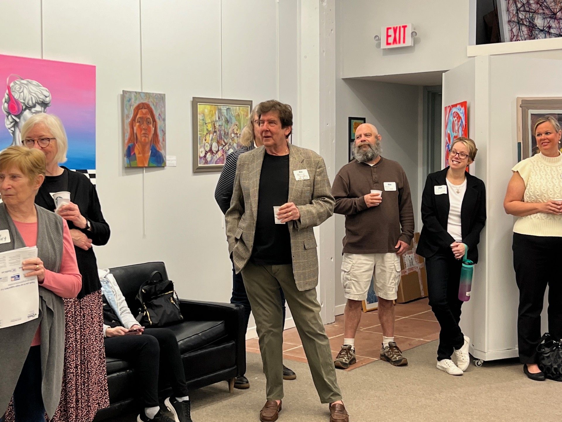 People in an art gallery, viewing paintings and socializing. Some are holding drinks, an exit sign visible.